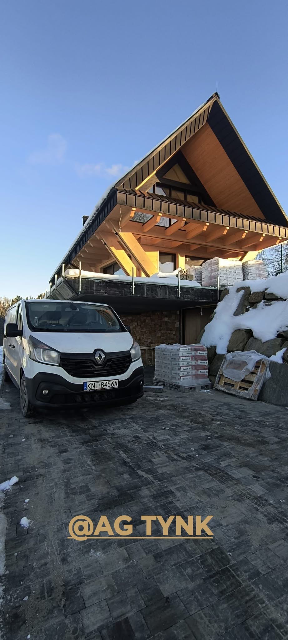 Nowoczesny dom z drewnianą konstrukcją, zaparkowany biały van i palety z materiałami budowlanymi na posesji. Widoczny fragment elewacji i kostka brukowa.