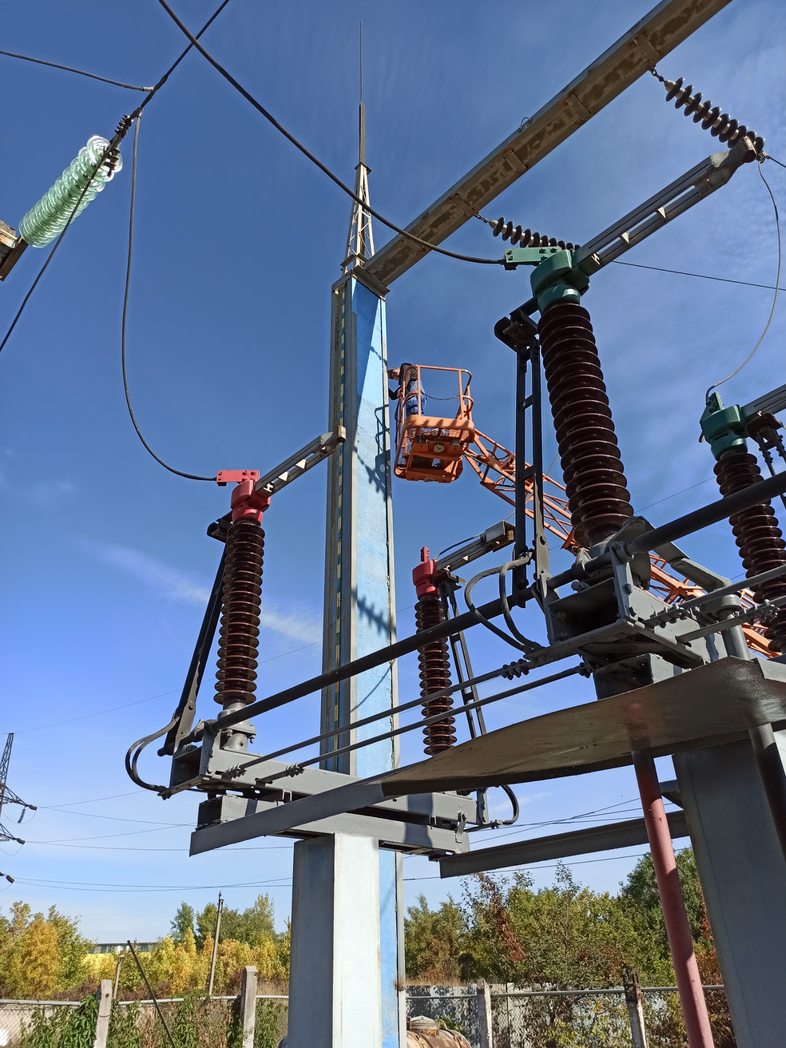Pracownik w podnośniku koszowym wykonuje prace na stacji transformatorowej wysokiego napięcia pod błękitnym niebem.