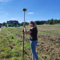 Blondynka w dżinsach obsługuje precyzyjny odbiornik GPS na tyczce geodezyjnej na polu z widocznym zielonym domem w tle.