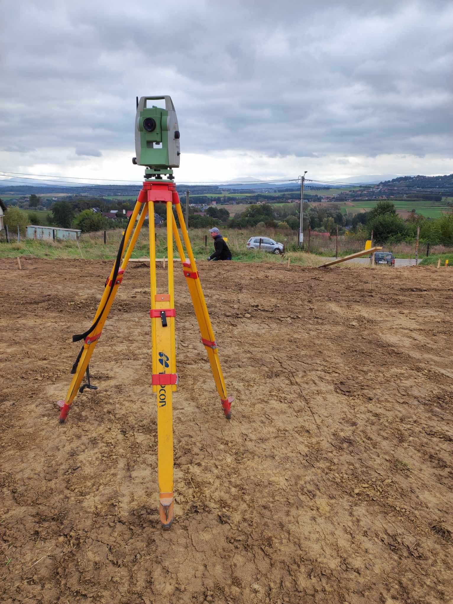 Żółty niwelator Topcon na trójnogu, ustawiony na nierównym, brązowym terenie budowy z widokiem na odległe zabudowania i pochmurne niebo.