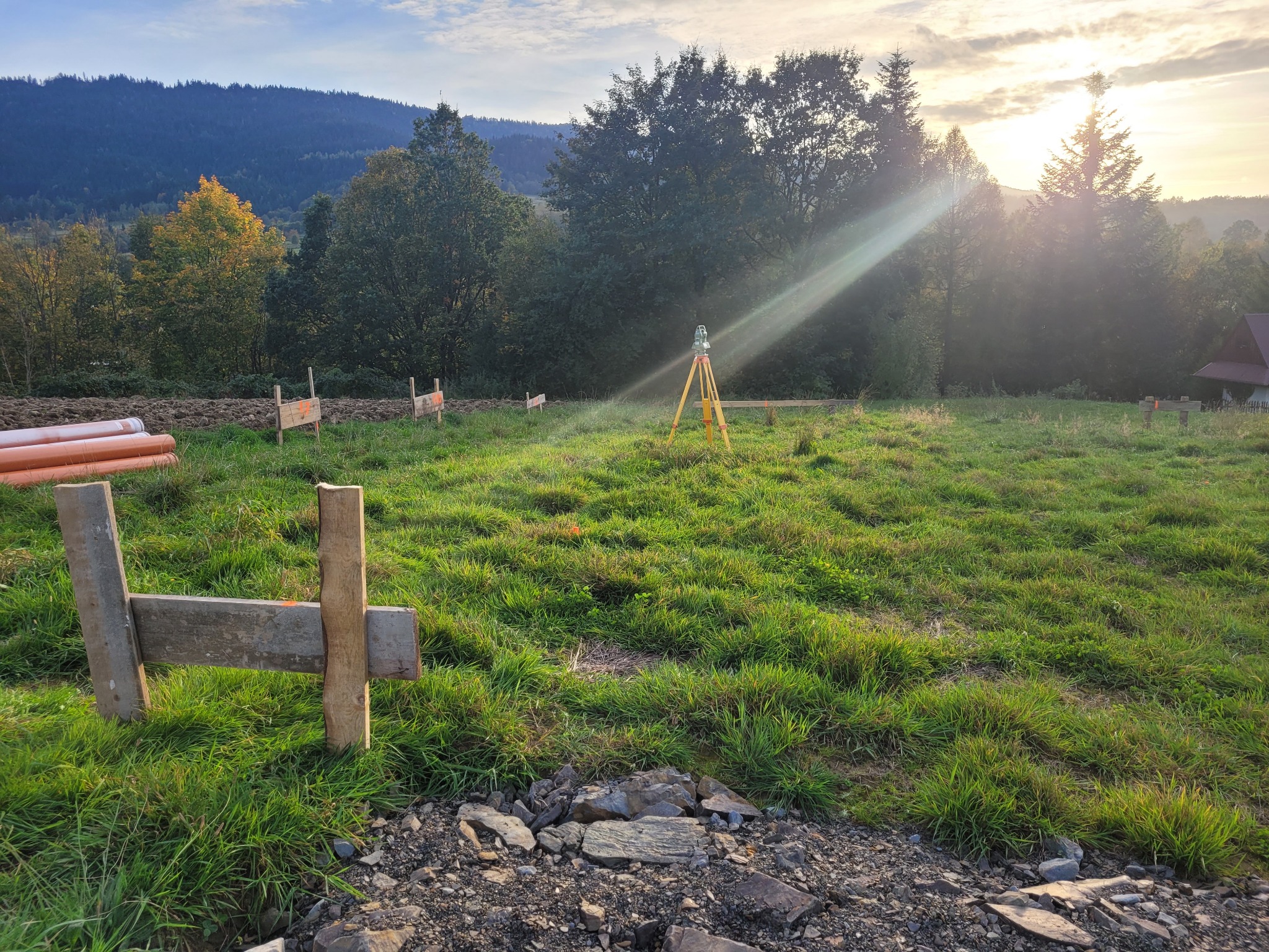 Żółty niwelator na trójnogu ustawiony na trawiastym terenie budowy z widokiem na zalesione wzgórza i wschodzące słońce, rury PCV i drewniane paliki wyznaczają obszar.