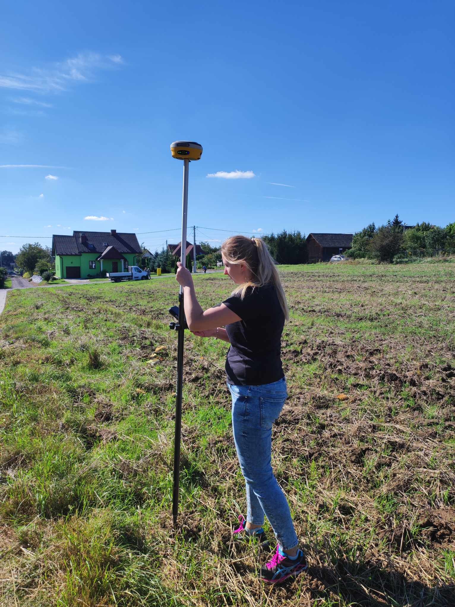 Blondynka w dżinsach obsługuje precyzyjny odbiornik GPS na tyczce geodezyjnej na polu z widocznym zielonym domem w tle.