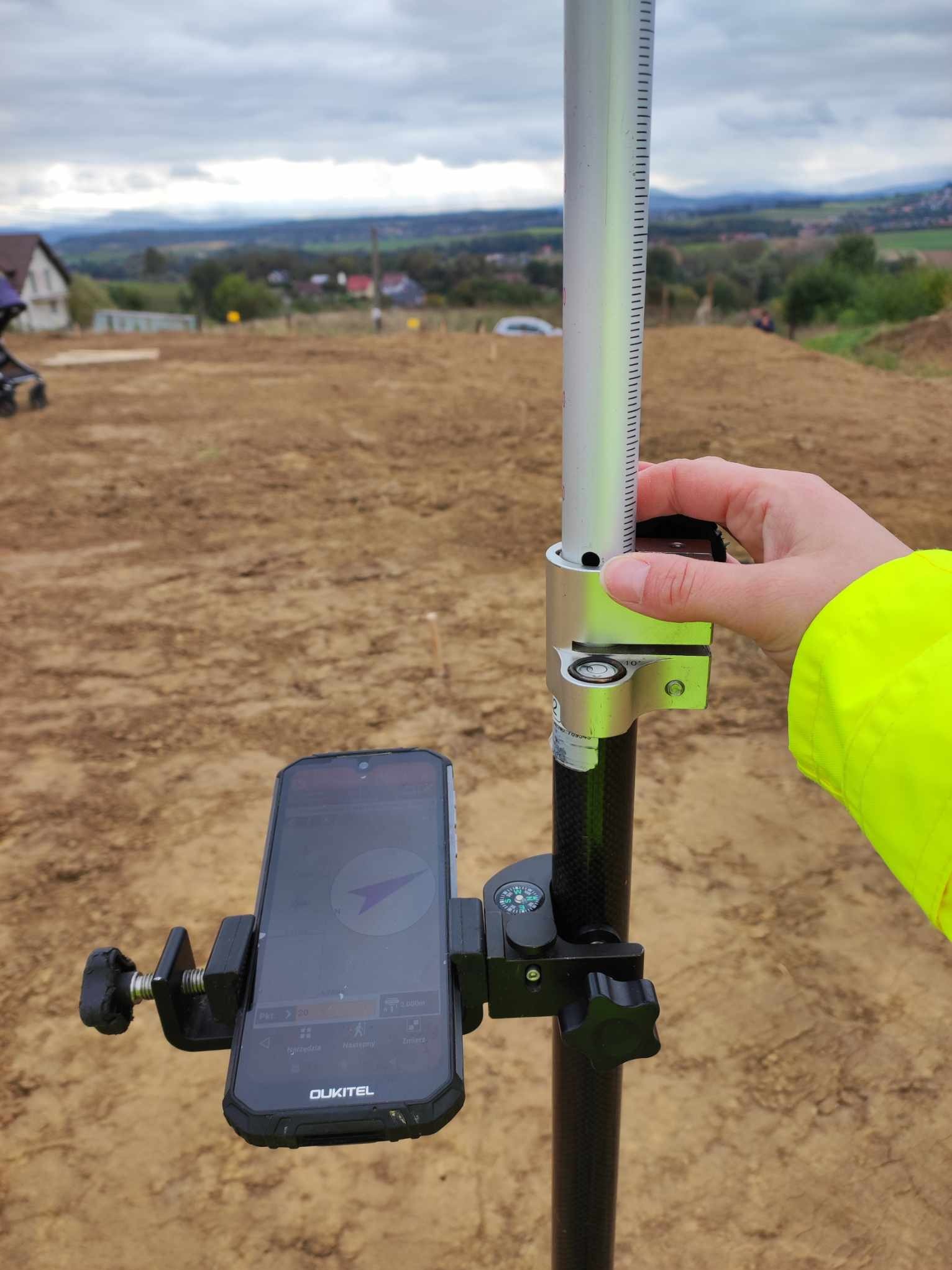 Pomiar gruntu w terenie: smartfon Oukitel zamocowany na tyczce geodezyjnej z kompasem, w tle widok na plac budowy i zabudowania.