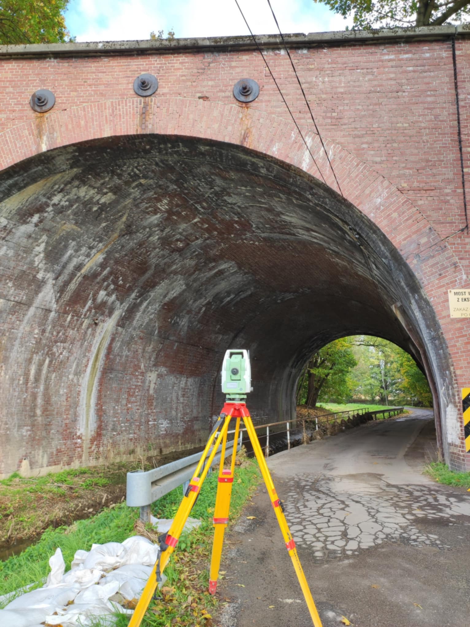 Zielony teodolit ustawiony na żółtym statywie przed ceglanym tunelem drogowym z widocznymi śladami wilgoci i zabrudzeń, w tle zieleń drzew i fragment drogi.