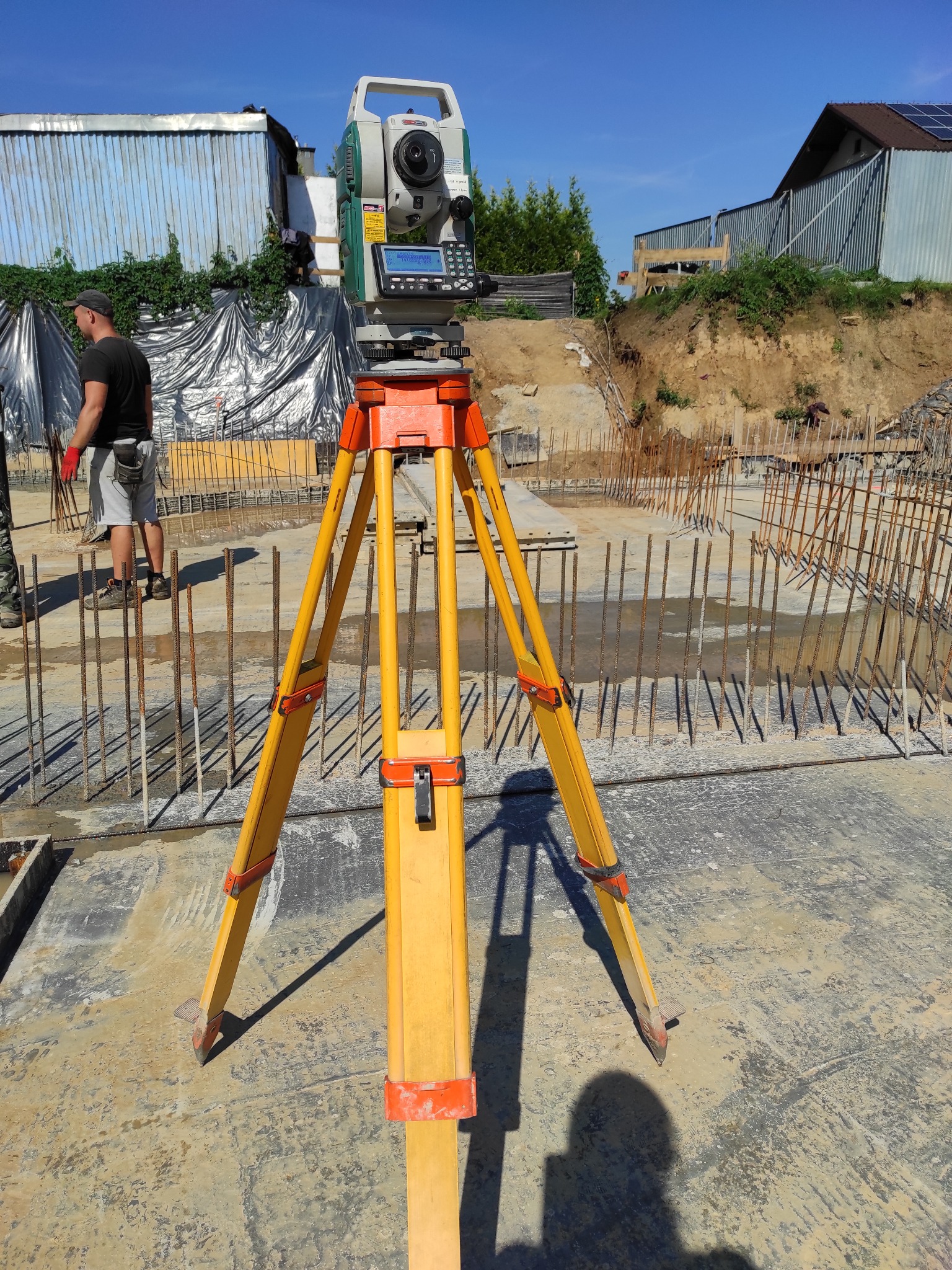 Pomarańczowo-żółty niwelator ustawiony na betonowym fundamencie z wystającymi prętami zbrojeniowymi, w tle pracownik w czerwonych rękawicach i budynek z blachy falistej.