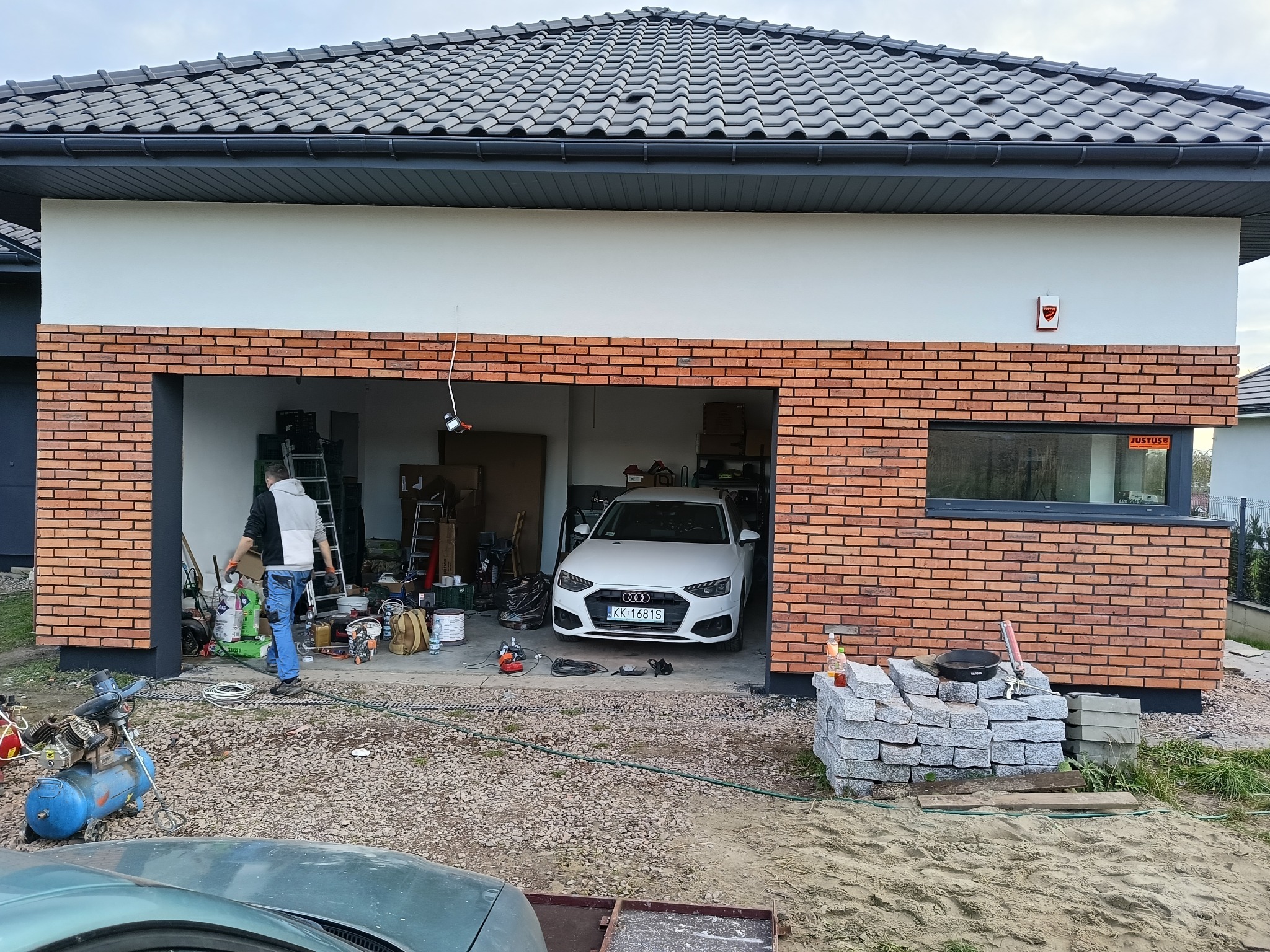 Nowoczesny dom z garażem w trakcie wykończenia elewacji z cegły klinkierowej. Widoczne narzędzia i materiały budowlane. Biały samochód w garażu.
