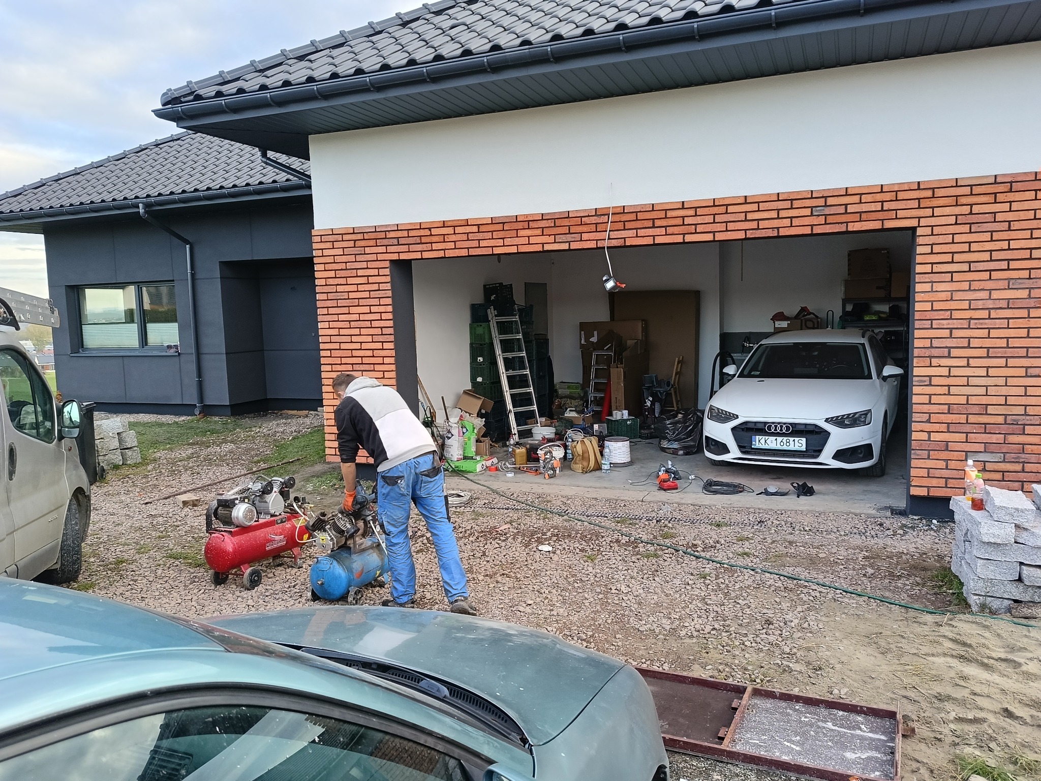 Prace murarskie przy garażu z cegły klinkierowej. Widoczne narzędzia, kompresory i częściowo samochód. W tle dom z szarą elewacją i grafitowym dachem.