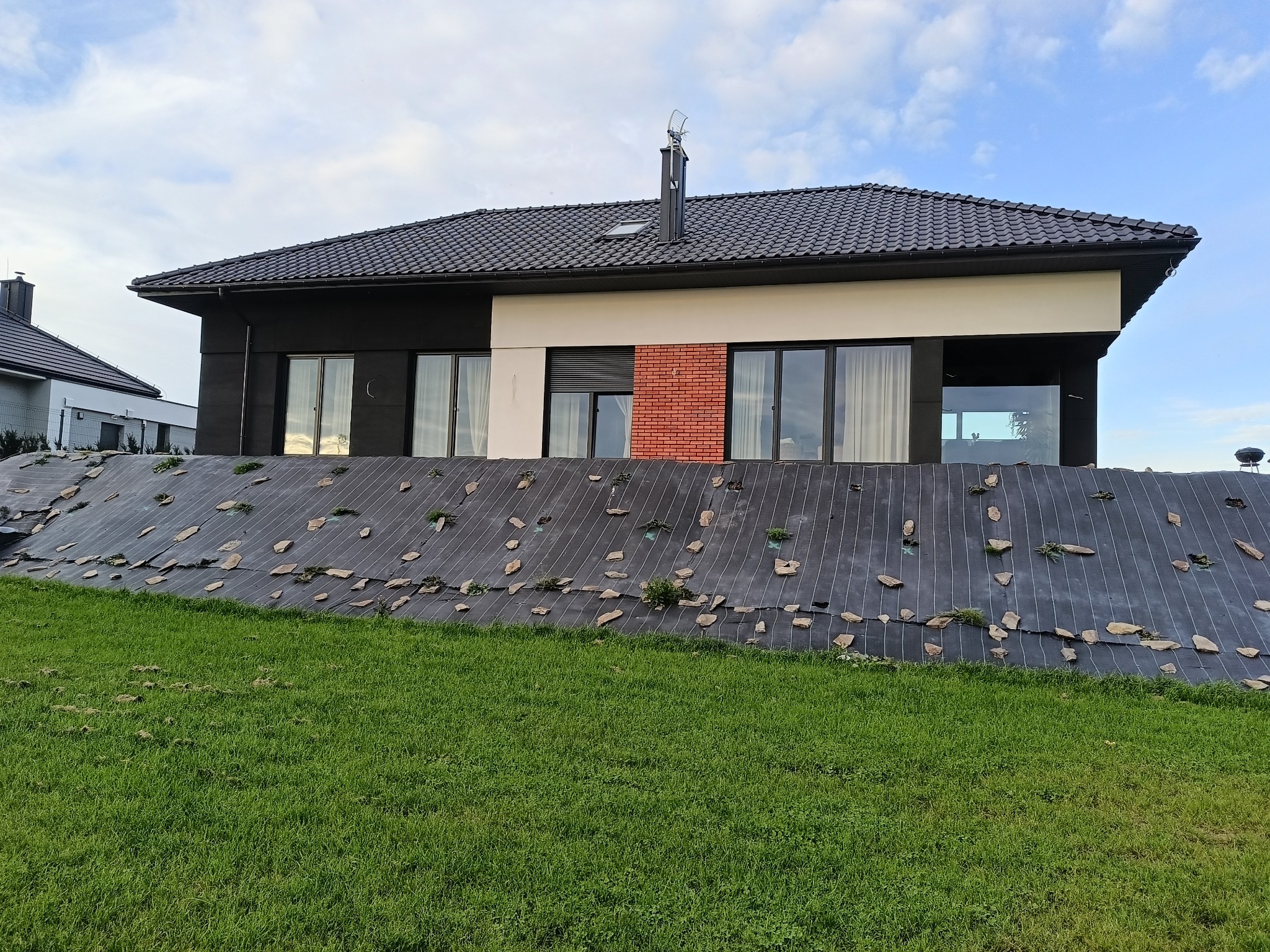 Nowoczesny dom jednorodzinny z ciemną dachówką i elewacją w odcieniach bieli, czerni i cegły. Uwagę zwraca zagospodarowanie terenu z użyciem agrowłókniny i kamieni.