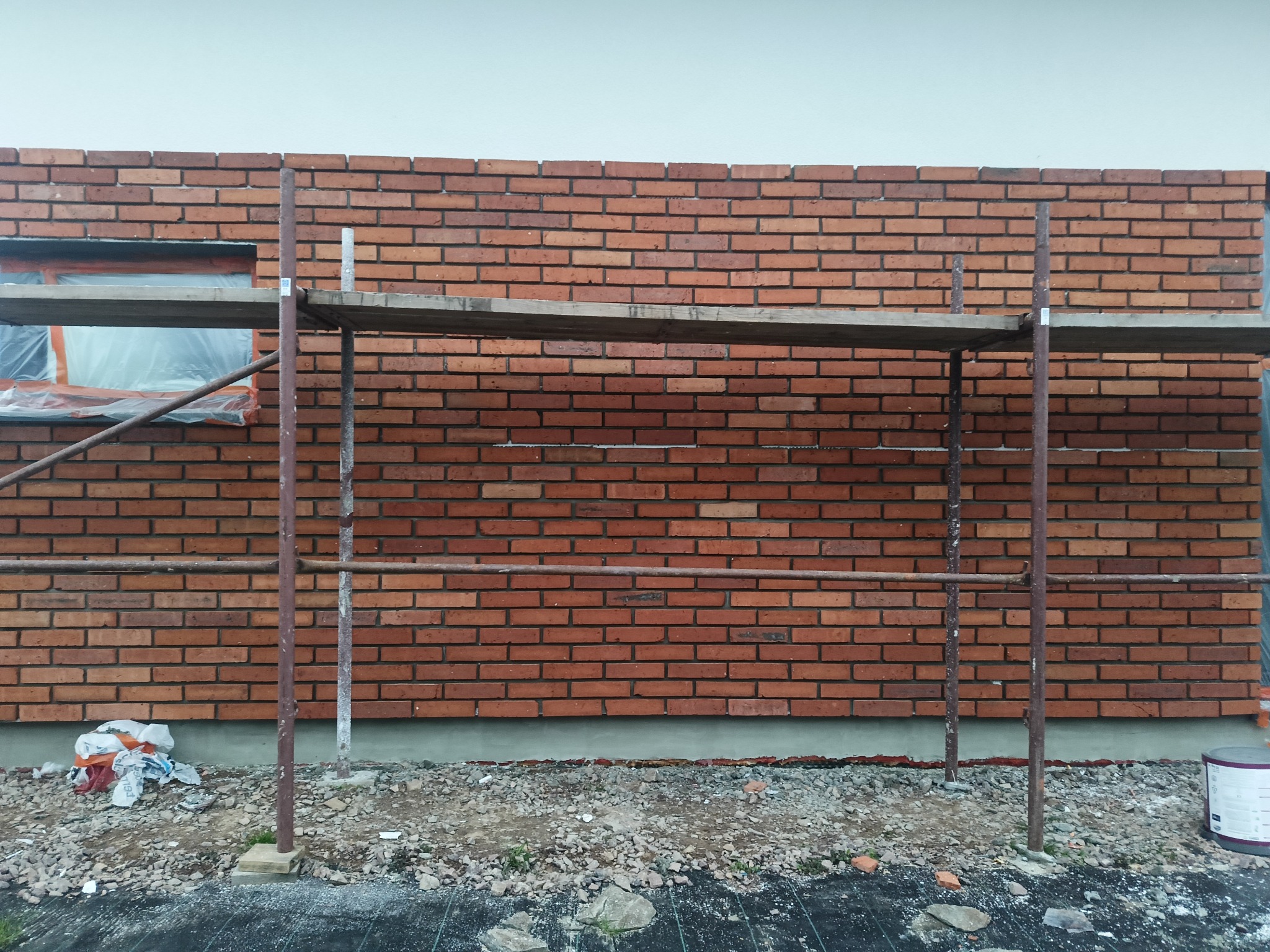 Murowana ściana z czerwonej cegły w trakcie budowy, widoczne rusztowanie i okno zabezpieczone folią. Fragment ściany z widocznymi spoinami i zaprawą.