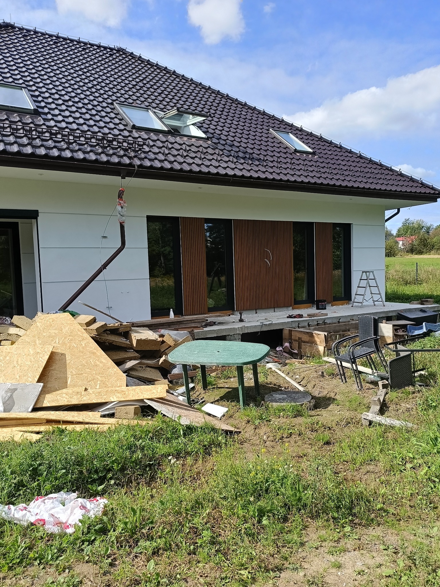 Dom jednorodzinny w trakcie prac wykończeniowych elewacji, widoczne materiały budowlane, okna z drewnianymi panelami i fragment dachu z oknami połaciowymi.