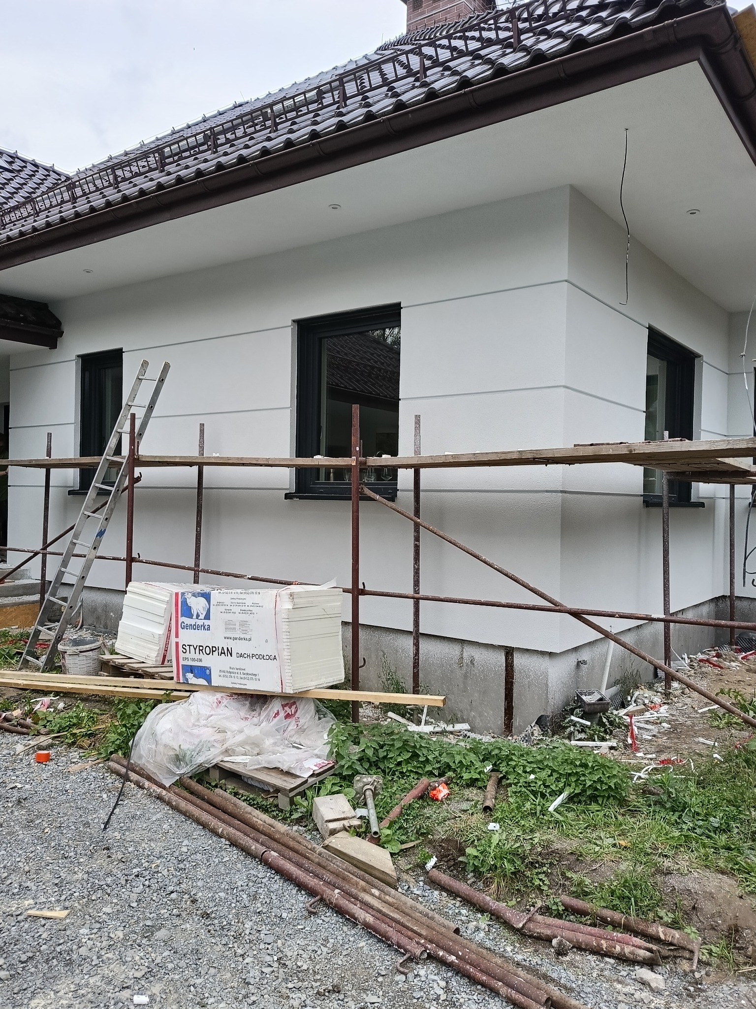 Ocieplony dom jednorodzinny w trakcie prac wykończeniowych elewacji, widoczne rusztowania, styropian dach-podłoga i drabina oparta o ścianę. Ujęcie z bliska.