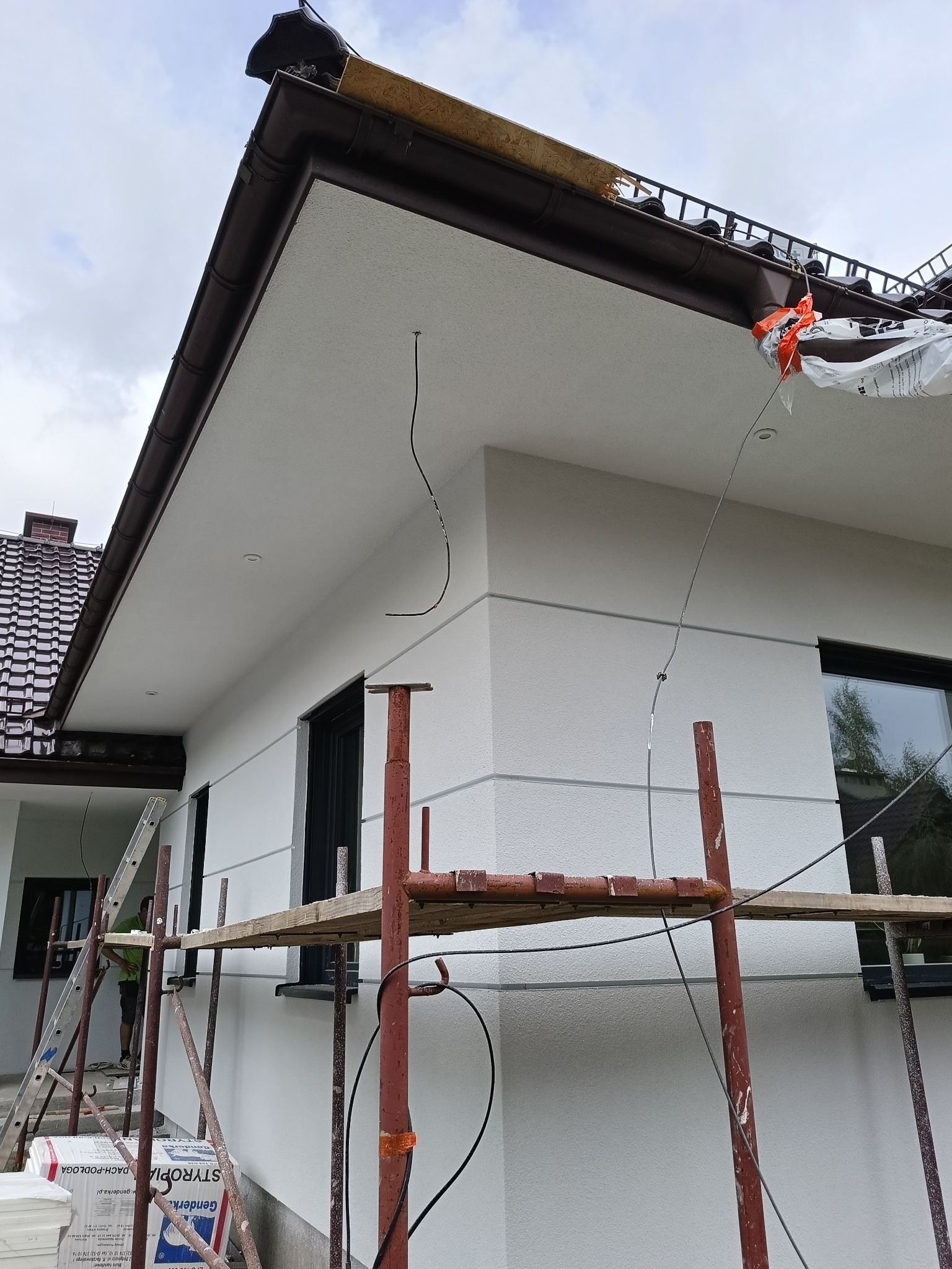 Biała elewacja domu jednorodzinnego z brązowymi rynnami, widoczna z dołu, z rusztowaniem i materiałami izolacyjnymi. Ukończona elewacja z widocznymi elementami wykończeniowymi.