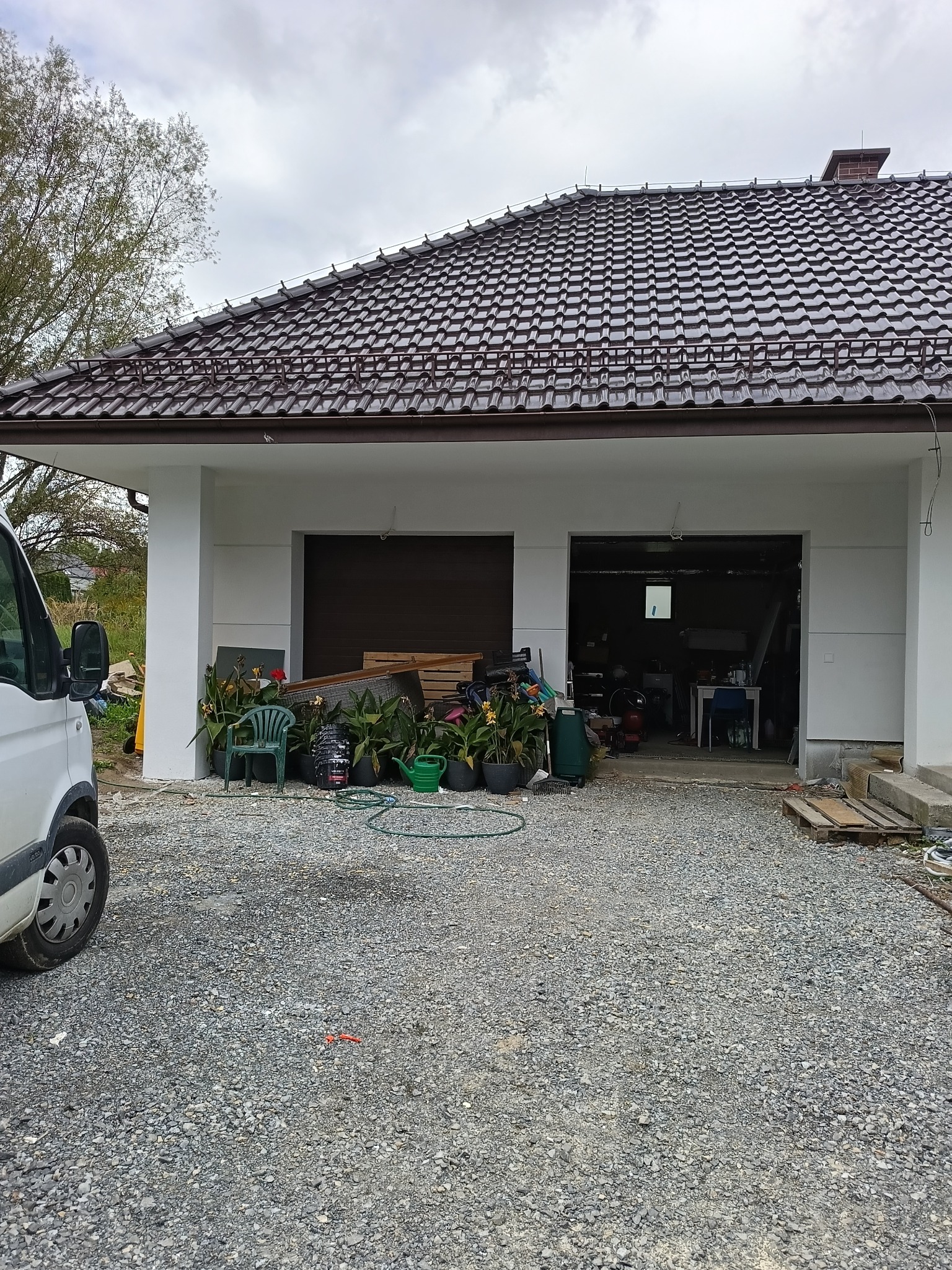 Dwustanowiskowy garaż z brązową bramą i świeżo pomalowaną elewacją na biało. Na podjeździe gruz i rośliny w donicach. Częściowo widoczny samochód dostawczy.