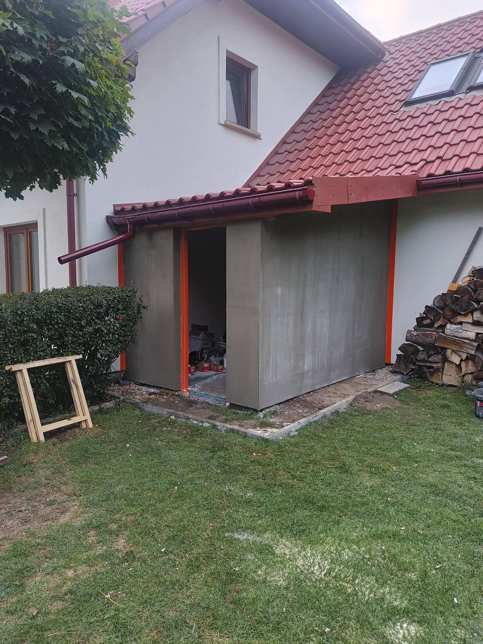 Szara, otynkowana dobudówka przy domu z czerwonym dachem i pomarańczowymi obramowaniami. Widoczne narzędzia wewnątrz. Na zewnątrz drewno i trawa.