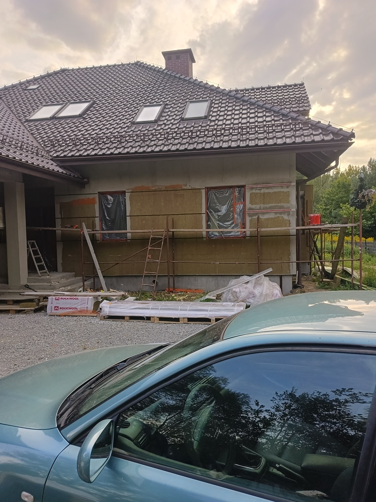 Dom w trakcie ocieplania: widoczne rusztowania, okna zabezpieczone folią, częściowo ocieplona ściana. W tle dach z oknami połaciowymi i fragment samochodu.