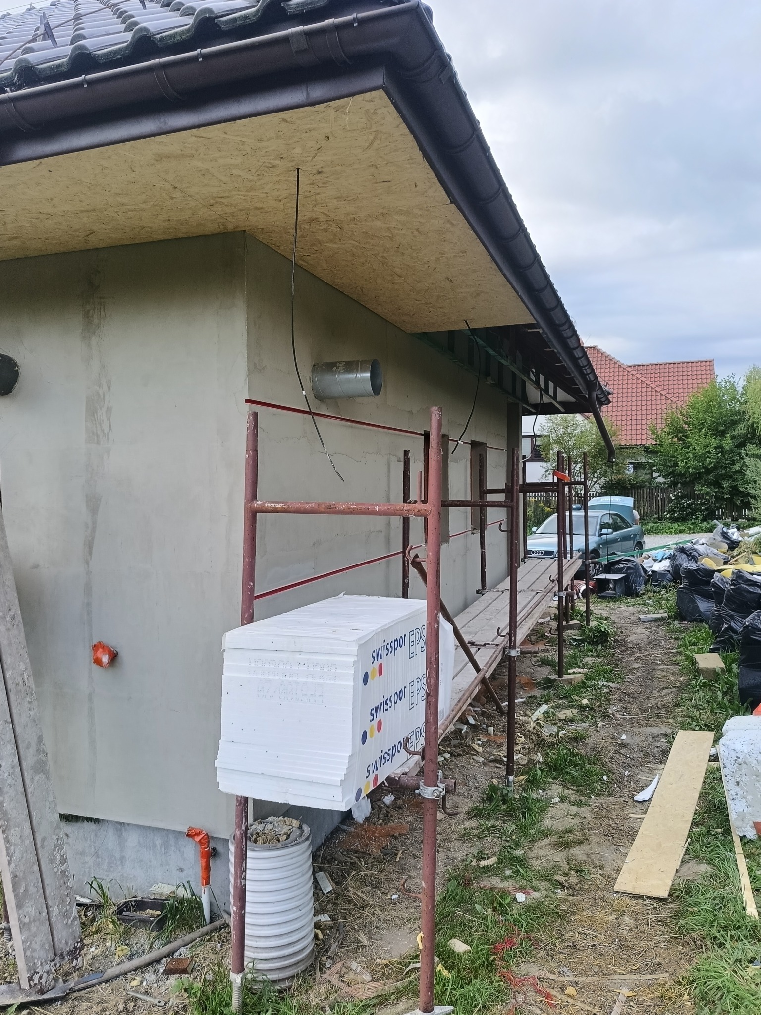 Budynek w trakcie ocieplania: styropian na rusztowaniu, surowa elewacja, widoczne elementy dachu i orynnowania. Prace w toku, otoczenie placu budowy.