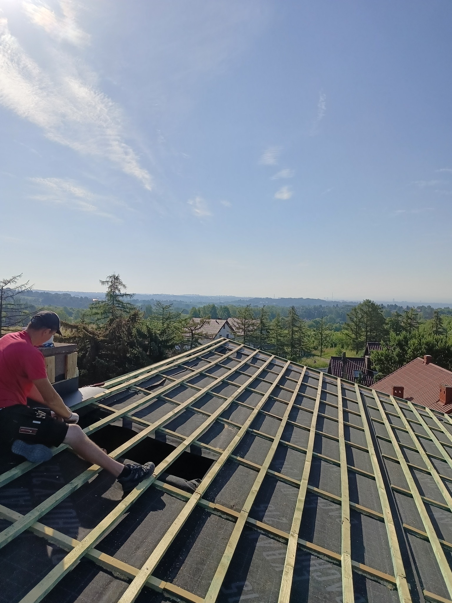 Dekarz montuje drewnianą konstrukcję dachu na czarnej membranie, z widokiem na zalesione wzgórza w tle, w słoneczny dzień. Widoczne narzędzia pracy.