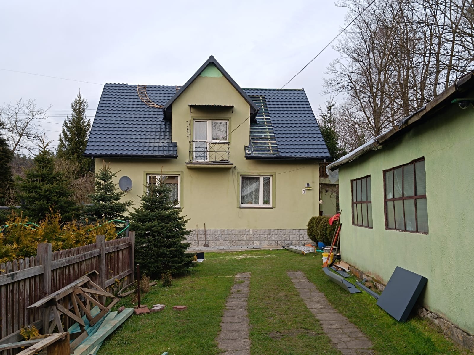 Dom z częściowo wymienionym, ciemnym dachem. Na dachu widoczne rusztowanie dekarskie. Elewacja w odcieniach zieleni, z balkonem i numerem domu '1'.