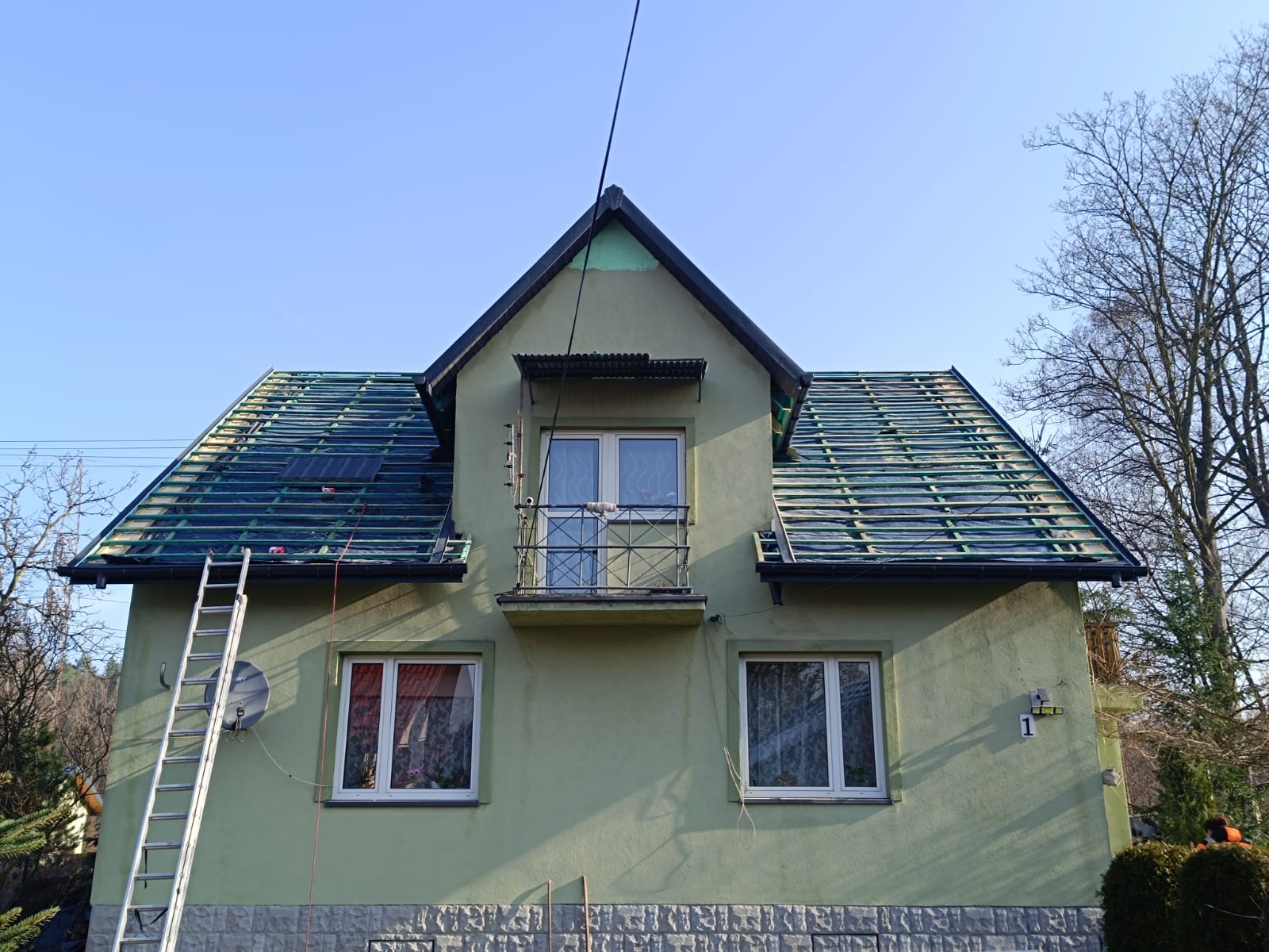 Dom z zieloną elewacją w trakcie krycia dachu. Widoczna drabina oparta o ścianę, przygotowane łaty pod dachówkę i fragment balkonu z metalową balustradą.