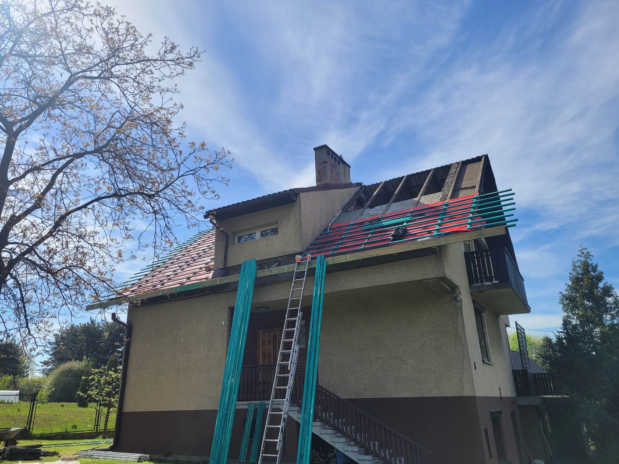 Dom z częściowo zdjętym dachem, widoczne elementy konstrukcyjne i przygotowane deski. Prace dekarskie w toku, drabina oparta o ścianę budynku.