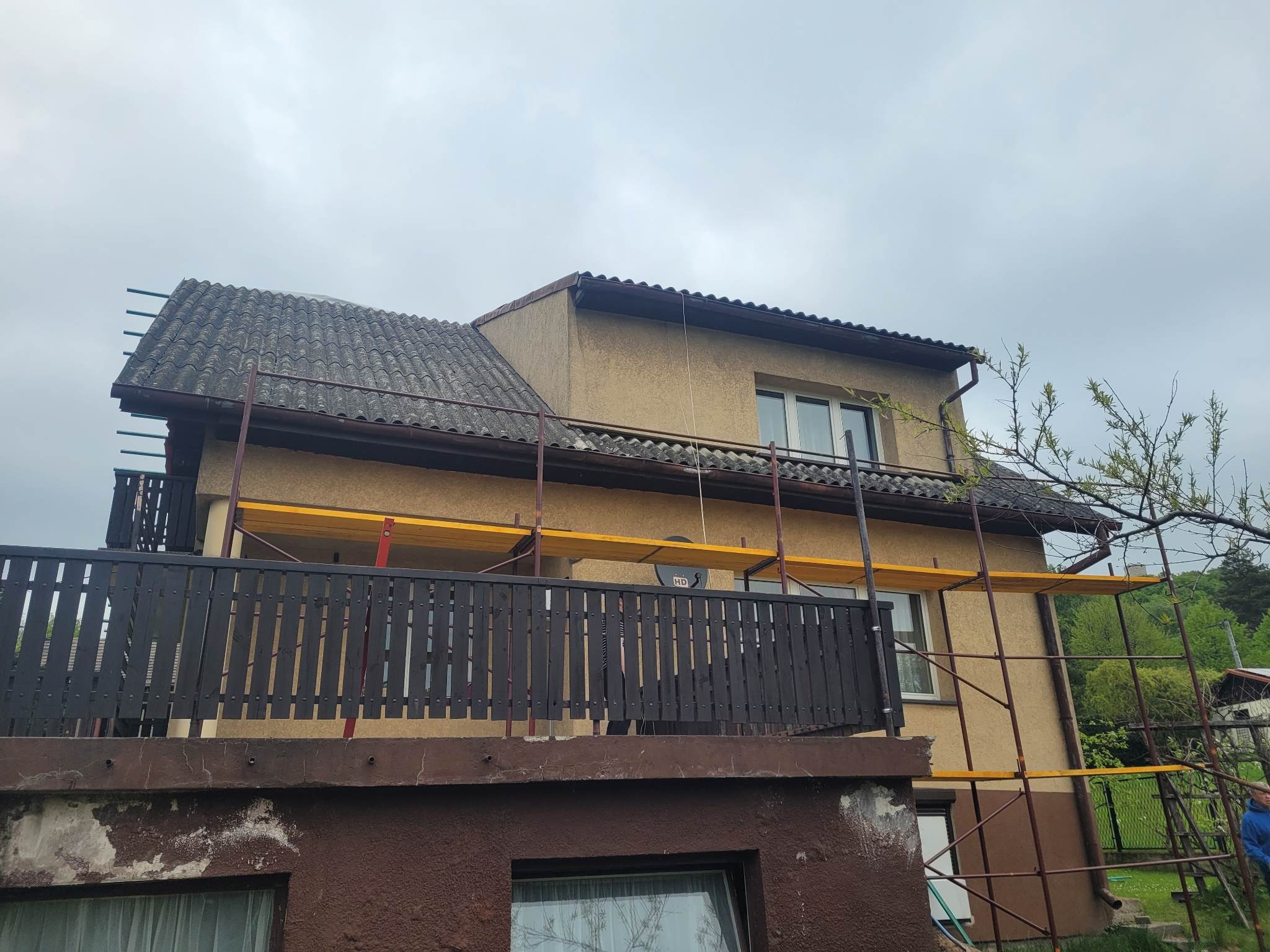Dom z elewacją w odcieniach beżu, otoczony rusztowaniem. Widoczny fragment dachu z szarej dachówki i ciemnobrązowa balustrada balkonu. Pochmurne niebo w tle.