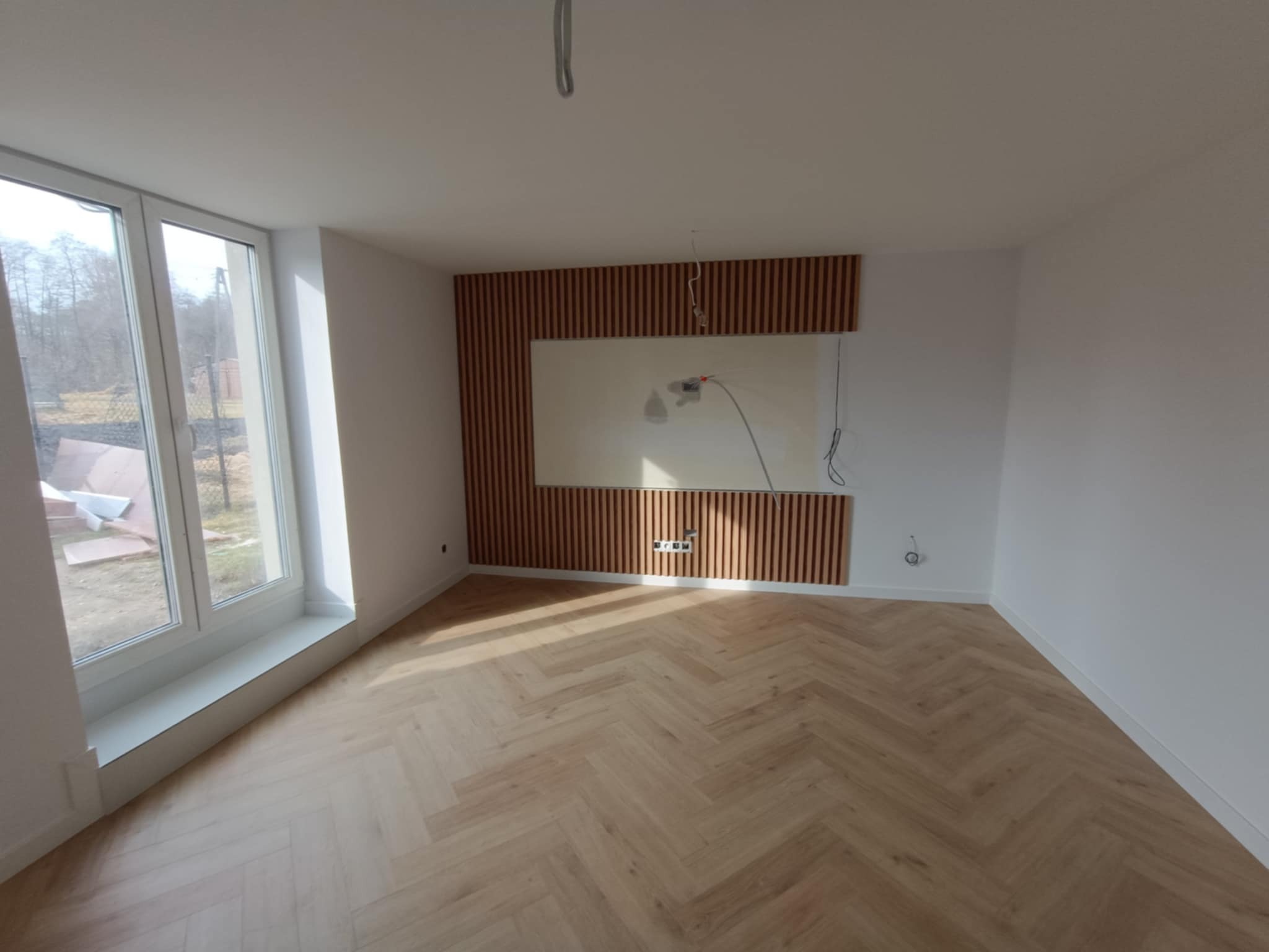 Jasne panele ułożone w jodełkę francuską w pokoju z dużym oknem. Na ścianie dekoracyjne lamele drewniane. Widoczne gniazdka elektryczne.