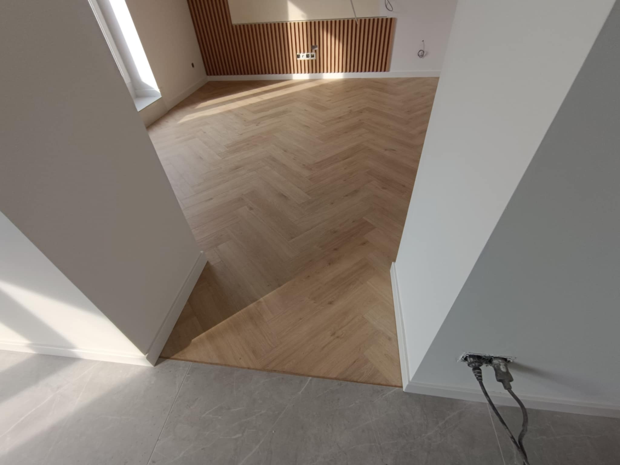 Ułożone panele podłogowe w jodełkę, widok z góry, jasne drewno, białe ściany, listwy przypodłogowe. Fragment szarej podłogi z płytek. Gniazdko elektryczne z kablami.