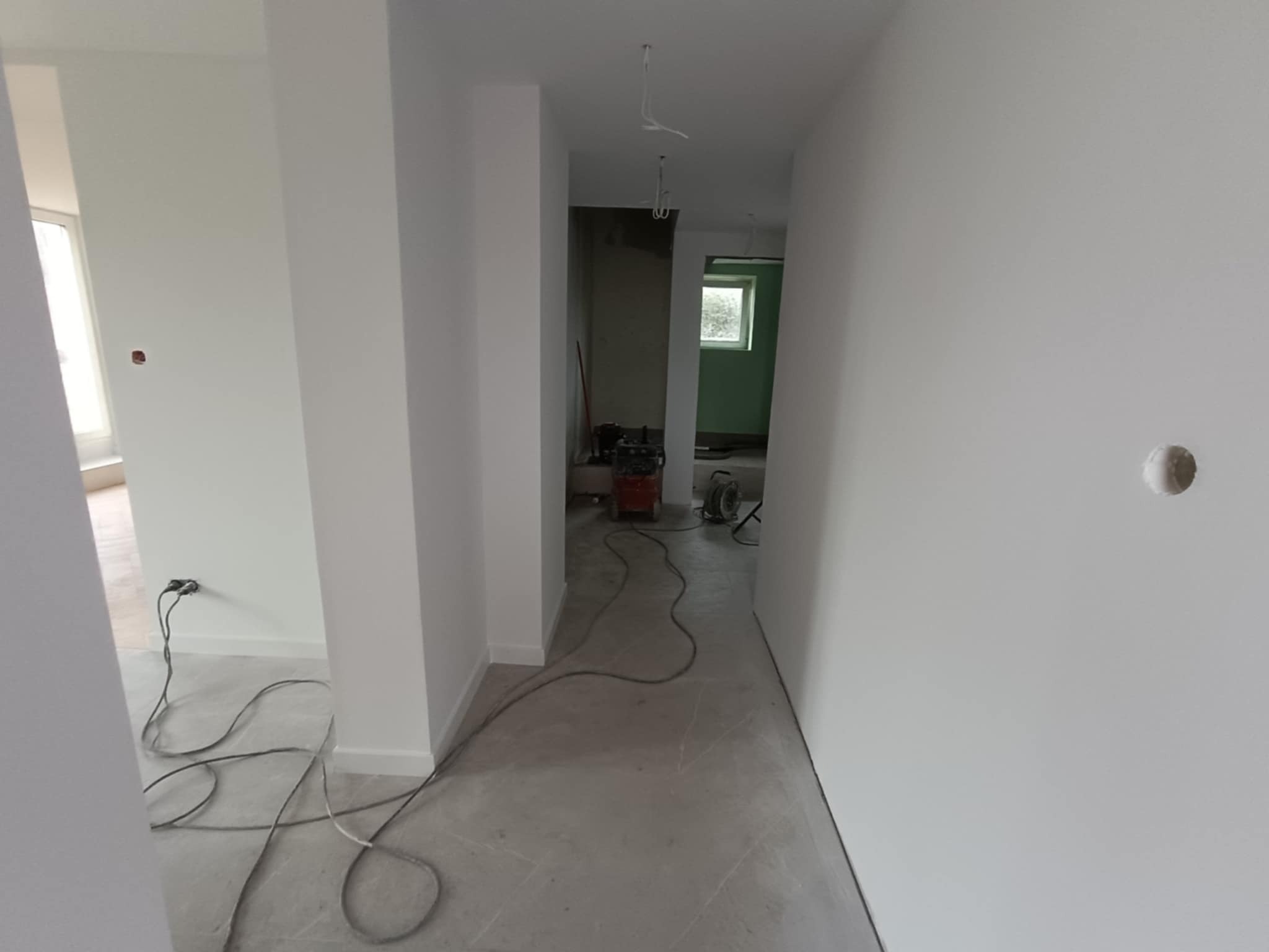 Wnętrze po malowaniu na biało, widoczne kable i sprzęt budowlany. Świeżo pomalowane ściany, surowa podłoga. Widok na dalsze pomieszczenia z oknem.
