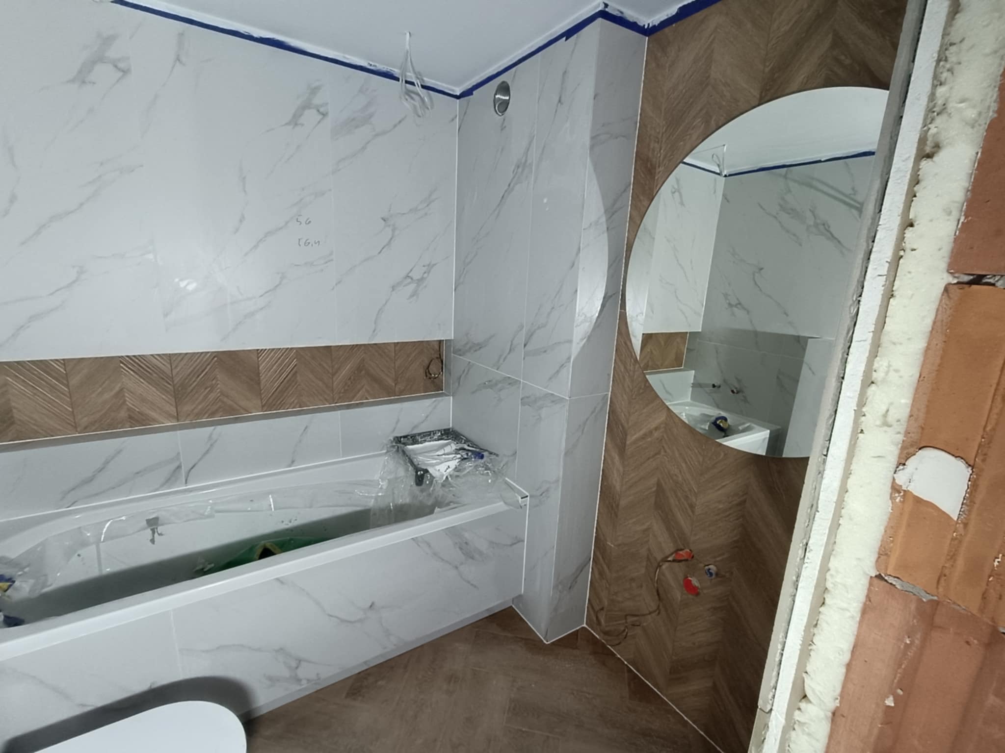 Remont łazienki: białe marmurowe płytki, drewnopodobne akcenty, okrągłe lustro. Widoczne zabezpieczona wanna i fragment ściany w trakcie wykończenia.