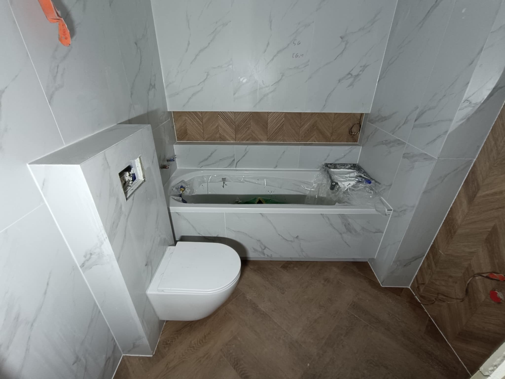 Wykończona łazienka z biało-szarymi płytkami imitującymi marmur, drewnopodobną mozaiką w niszy nad wanną i podłogą w jodełkę. Wisząca toaleta z zabudową.