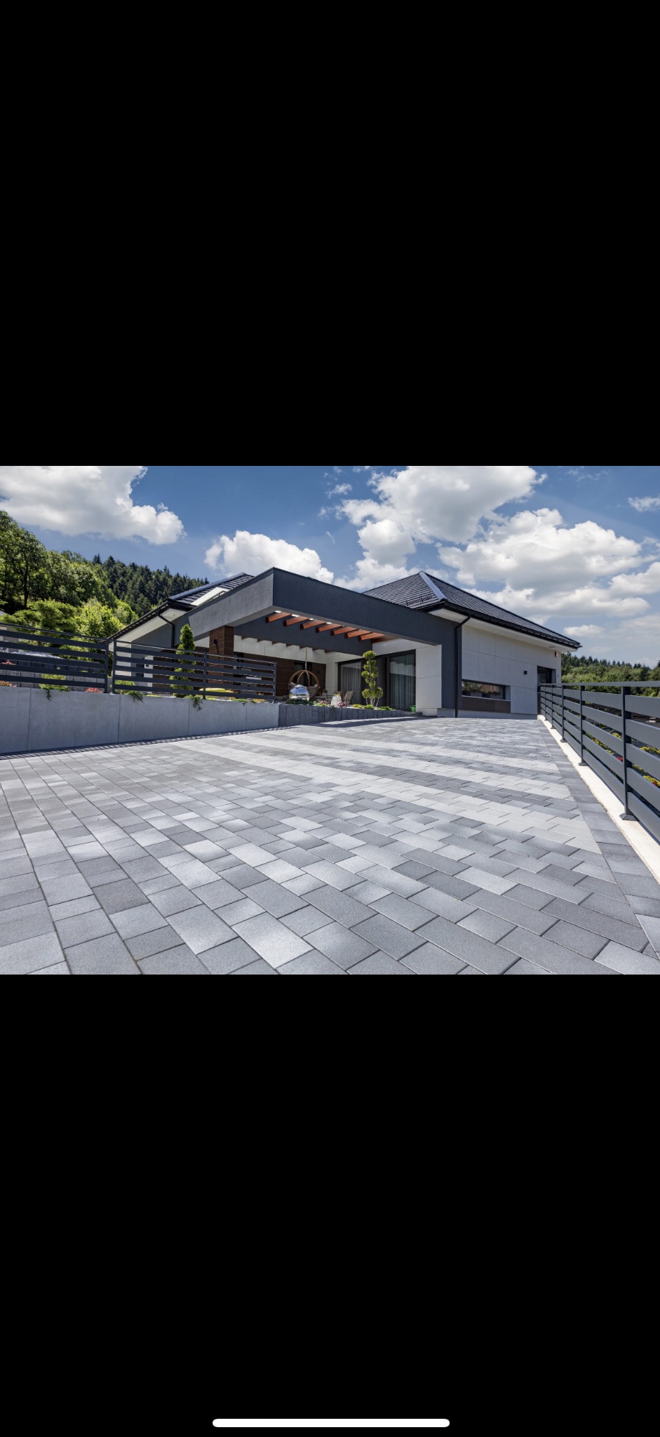 Nowoczesny dom z podjazdem wyłożonym szarą kostką brukową w geometryczny wzór. Widok na budynek z pergolą i elementami zieleni. Całość w słoneczny dzień.