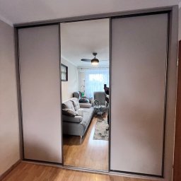 MM Meble Michał Misiło - Trzydrzwiowa szafa z przesuwnymi drzwiami, środkowe drzwi lustrzane odbijają wnętrze salonu z szarą sofą i dywanem.