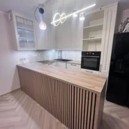 Nowoczesna kuchnia z białymi szafkami i drewnianym blatem wyspowym z pionowych listew, oświetlona designerskimi lampami wiszącymi.