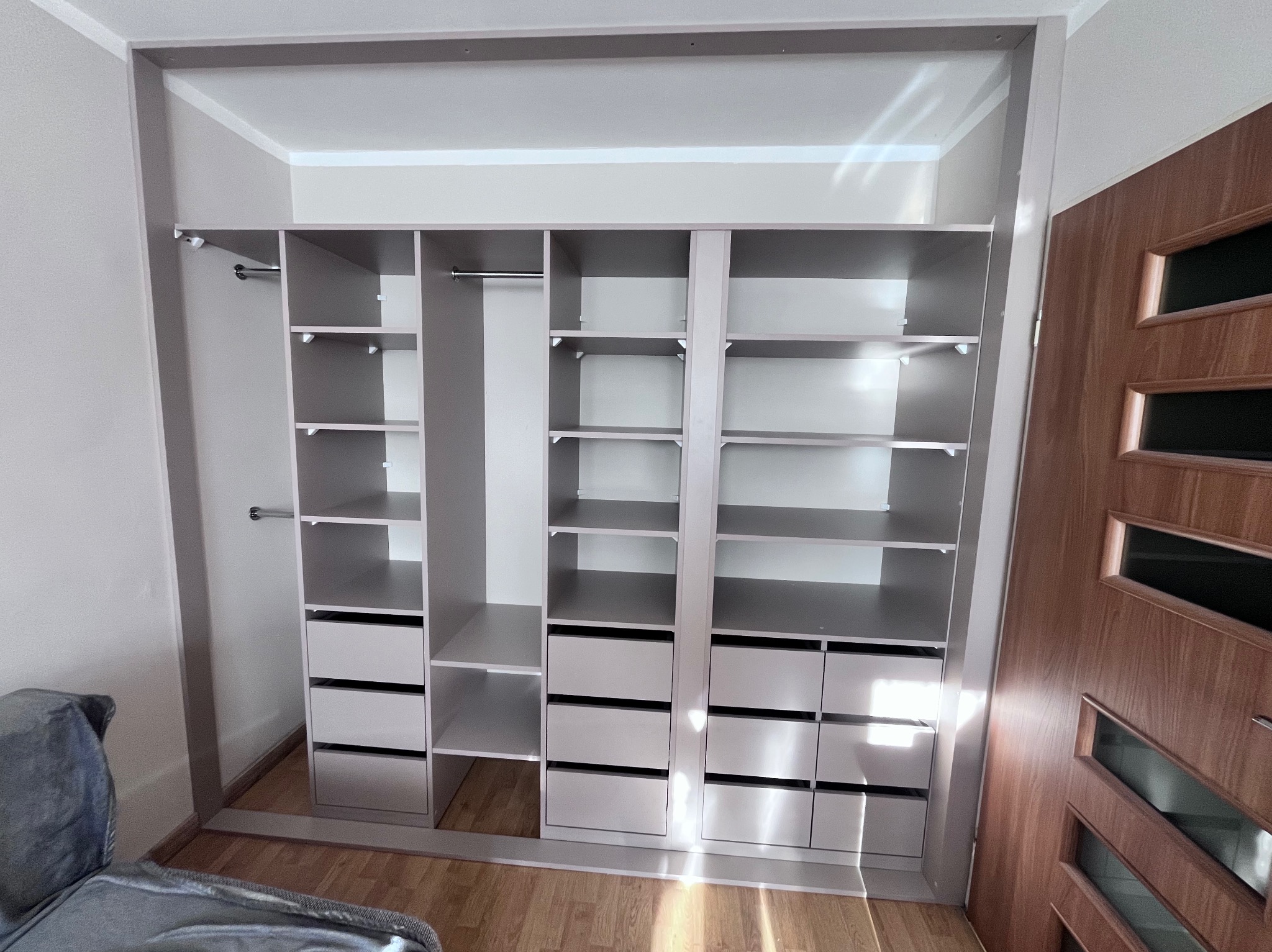 Jasnoszara zabudowa wnękowa z półkami i szufladami, widoczna z perspektywy pokoju z drewnianą podłogą i częścią sofy po lewej stronie, obok drzwi z przeszkleniami.