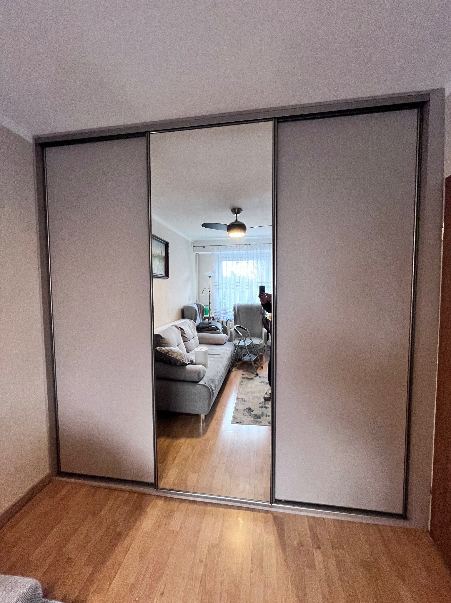 Trzydrzwiowa szafa z przesuwnymi drzwiami, środkowe drzwi lustrzane odbijają wnętrze salonu z szarą sofą i dywanem.