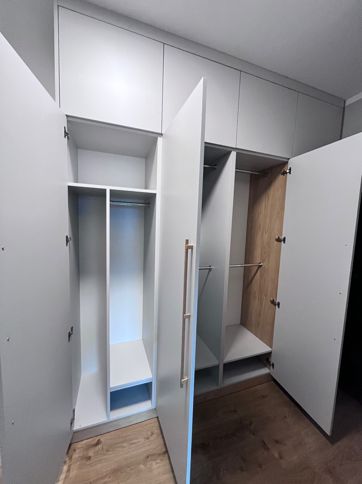 Jasnoszara zabudowa meblowa od podłogi do sufitu w Olsztynie, z otwartymi drzwiami ukazującymi półki i drążki na ubrania, drewniane panele wewnątrz szafy po prawej stronie.