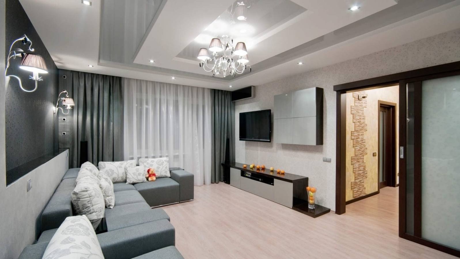 Nowoczesny salon z szarą sofą narożną, dekoracyjnymi poduszkami, telewizorem na ścianie, przesuwnymi drzwiami i oświetleniem sufitowym.