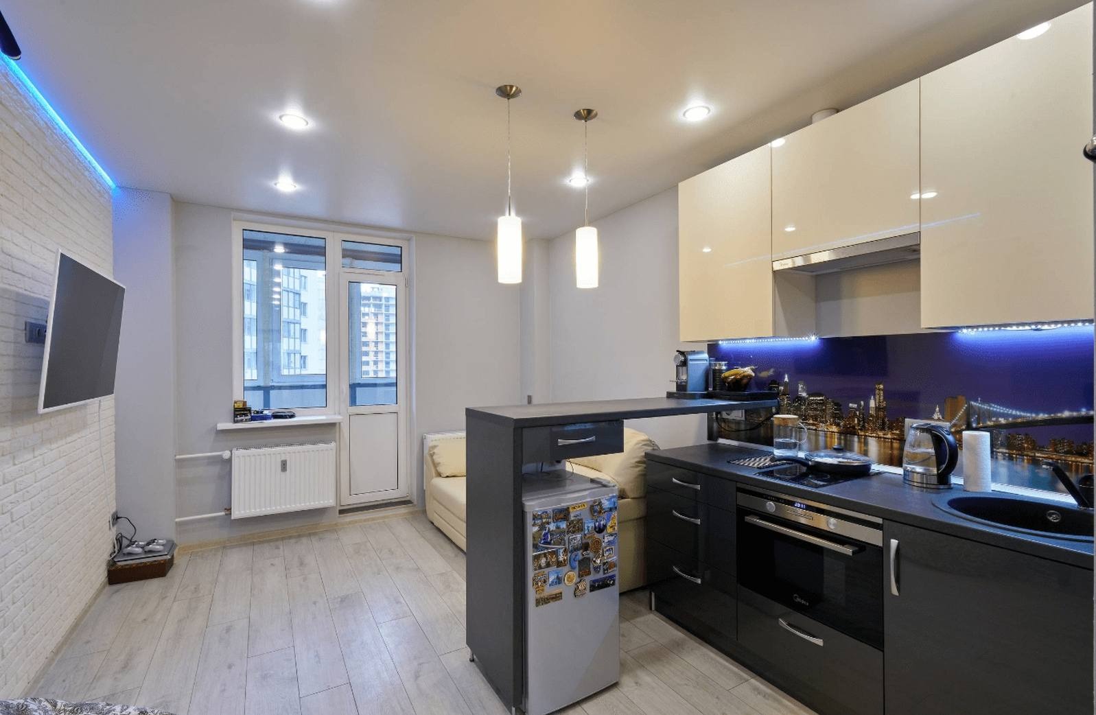 Nowoczesna kuchnia w mieszkaniu z aneksem, biało-czarne szafki, podświetlany panel z motywem miasta, lodówka z magnesami, drewniana podłoga, oświetlenie LED.