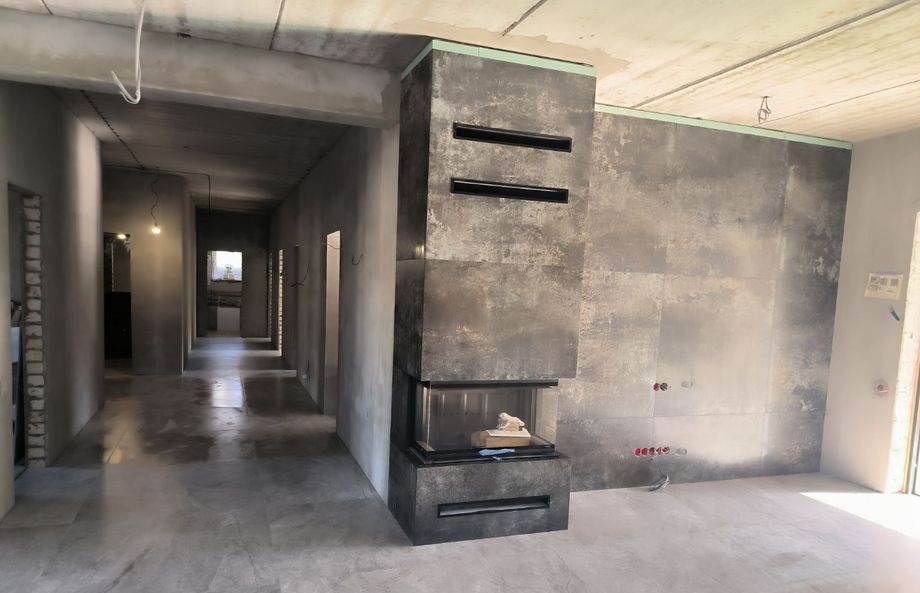 Surowe wnętrze w trakcie prac wykończeniowych: widoczny kominek obłożony płytami imitującymi beton, ściany przygotowane pod malowanie, surowy betonowy sufit i posadzka, oraz instalacje elektryczne...