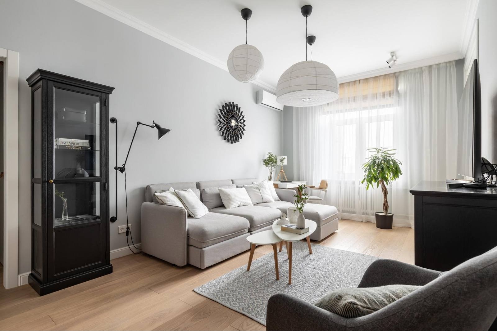 Jasne wnętrze salonu z szarą sofą, białymi lampami kulami i drewnianą podłogą, akcentowane czarną witryną i dekoracyjnym zegarem słonecznym.