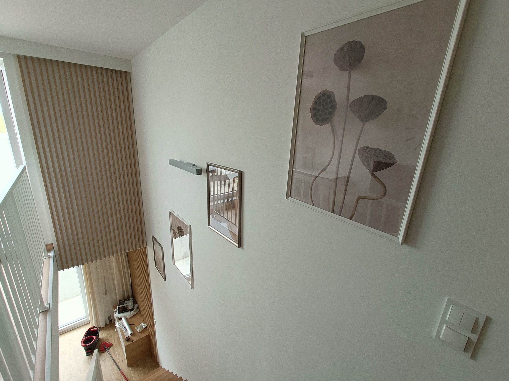 Widok z góry na klatkę schodową po odświeżeniu, z pomalowanymi na biało ścianami, drewnianymi panelami dekoracyjnymi na ścianie przy oknie i obrazami w ramkach.