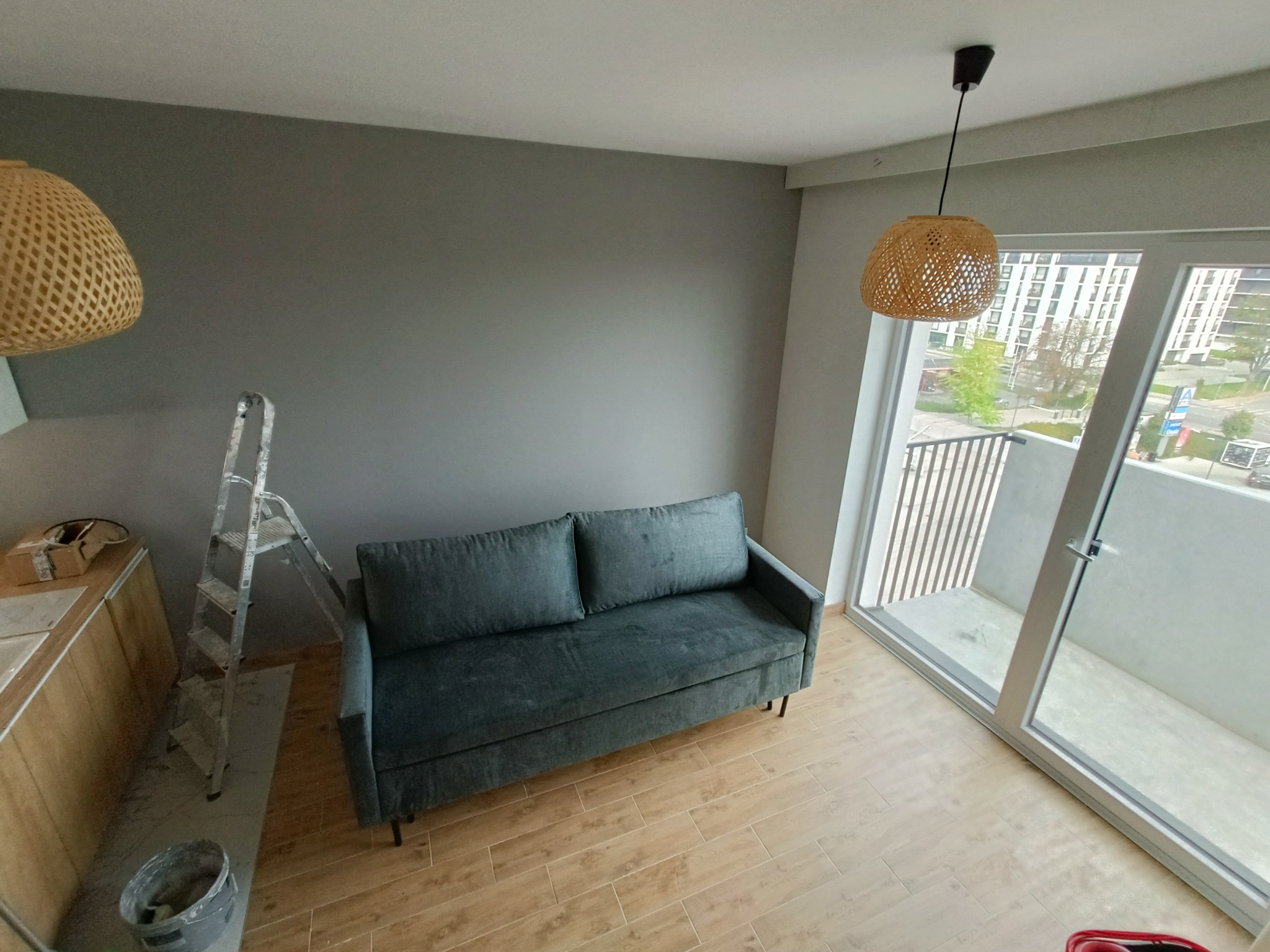 Szara ściana w nowym mieszkaniu po malowaniu, z drewnianą podłogą, kanapą w odcieniach szarości, drabina malarska, lampy z wikliny pod sufitem i balkonem z widokiem na miasto.