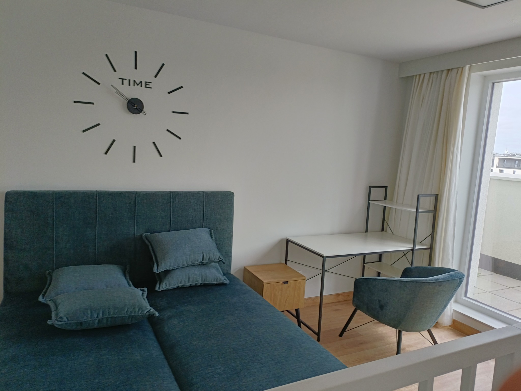 Nowoczesna sypialnia z łóżkiem z turkusowym zagłówkiem, biurkiem, krzesłem i zegarem ściennym, widok na balkon.