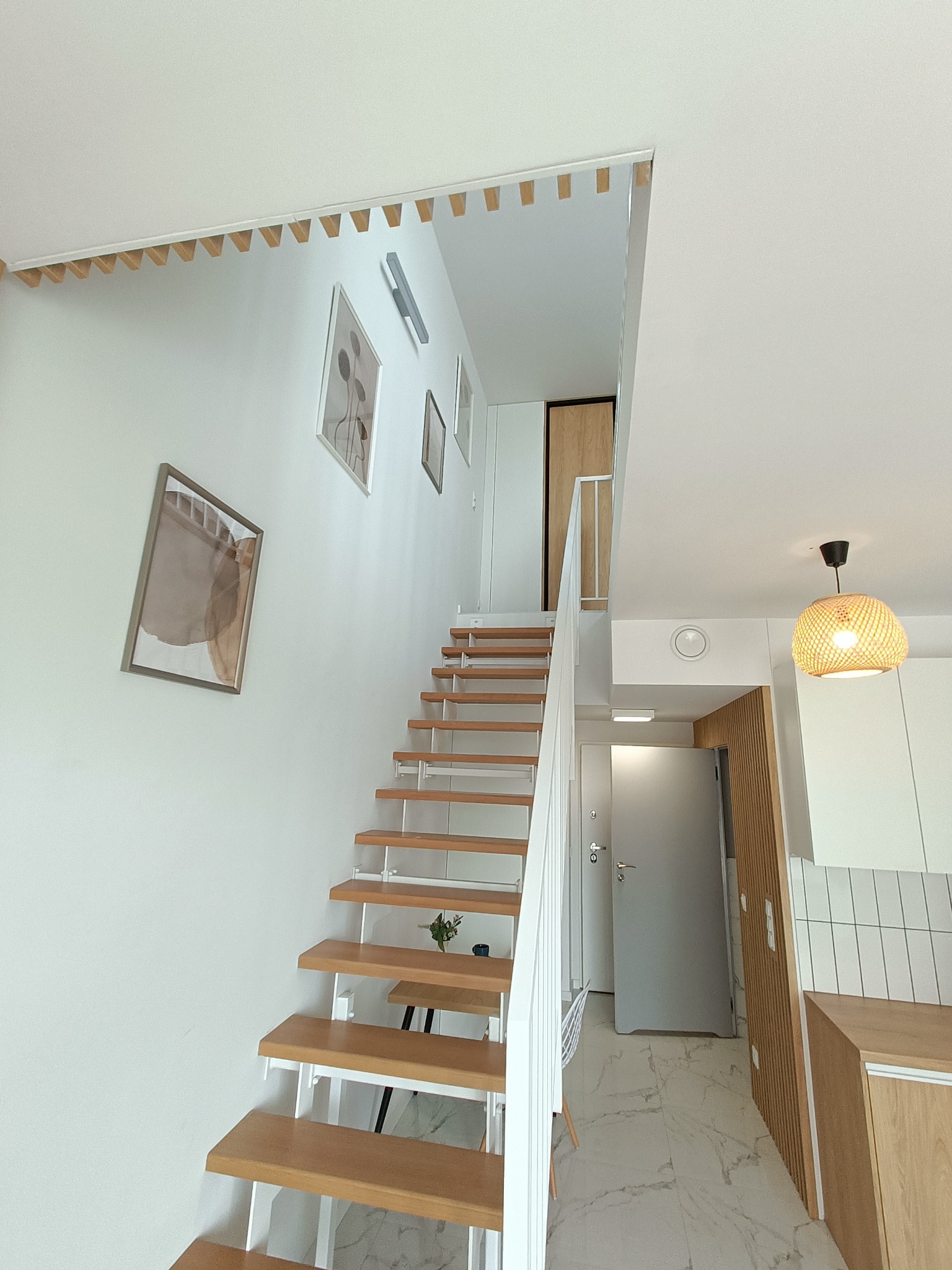 Nowoczesne schody drewniane z białymi wspornikami w jasnym wnętrzu, minimalistyczne obrazy na ścianie, dekoracyjne wykończenie sufitu.