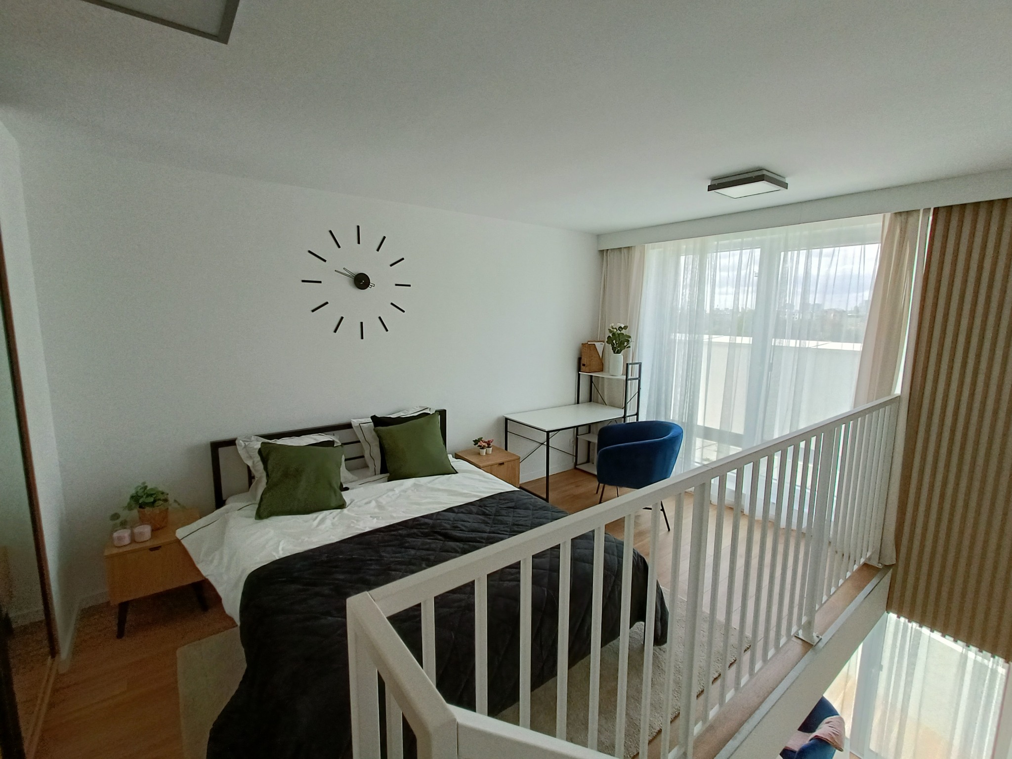 Nowoczesna sypialnia na antresoli z białymi ścianami, minimalistycznym zegarem, łóżkiem z zielonymi poduszkami, biurkiem i niebieskim krzesłem, widok z góry przez białą barierkę.