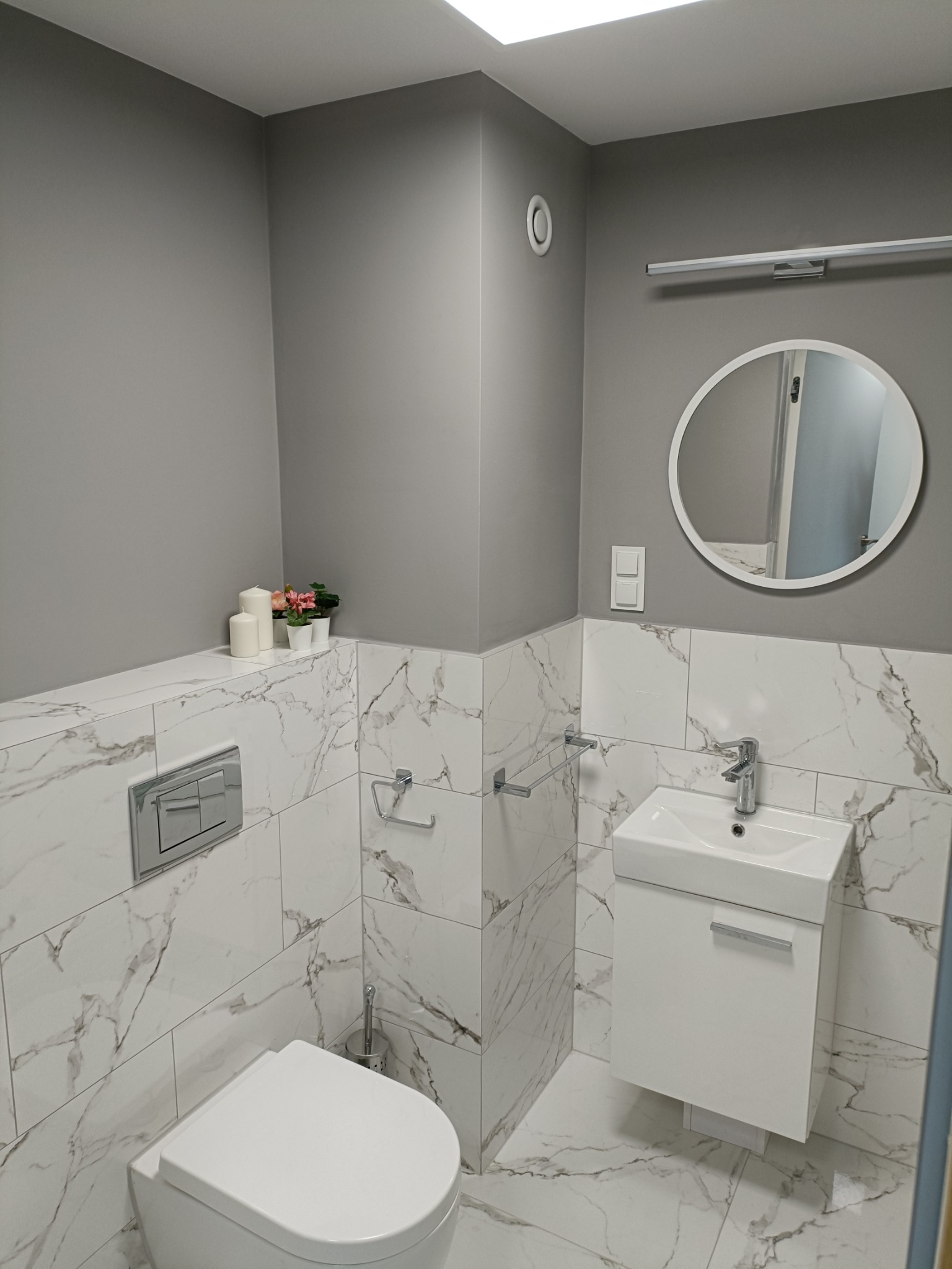 Nowoczesna łazienka w szaro-białej kolorystyce z marmurowymi płytkami, wisząca toaleta, umywalka z szafką, okrągłe lustro i dekoracyjne świece z kwiatami na półce.