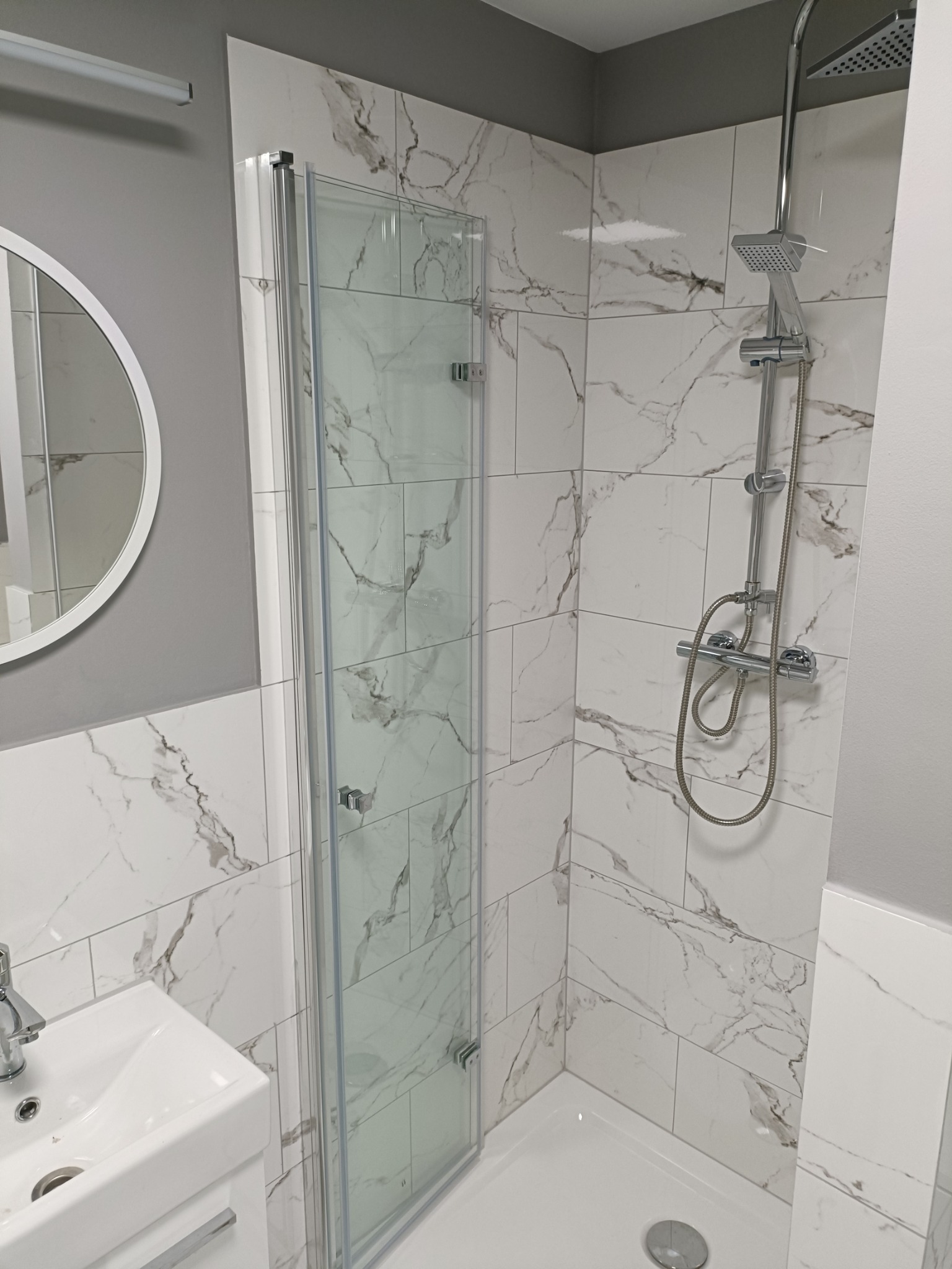 Nowoczesna kabina prysznicowa z deszczownicą i panelami imitującymi marmur, umywalka z baterią w tle, oświetlenie sufitowe.