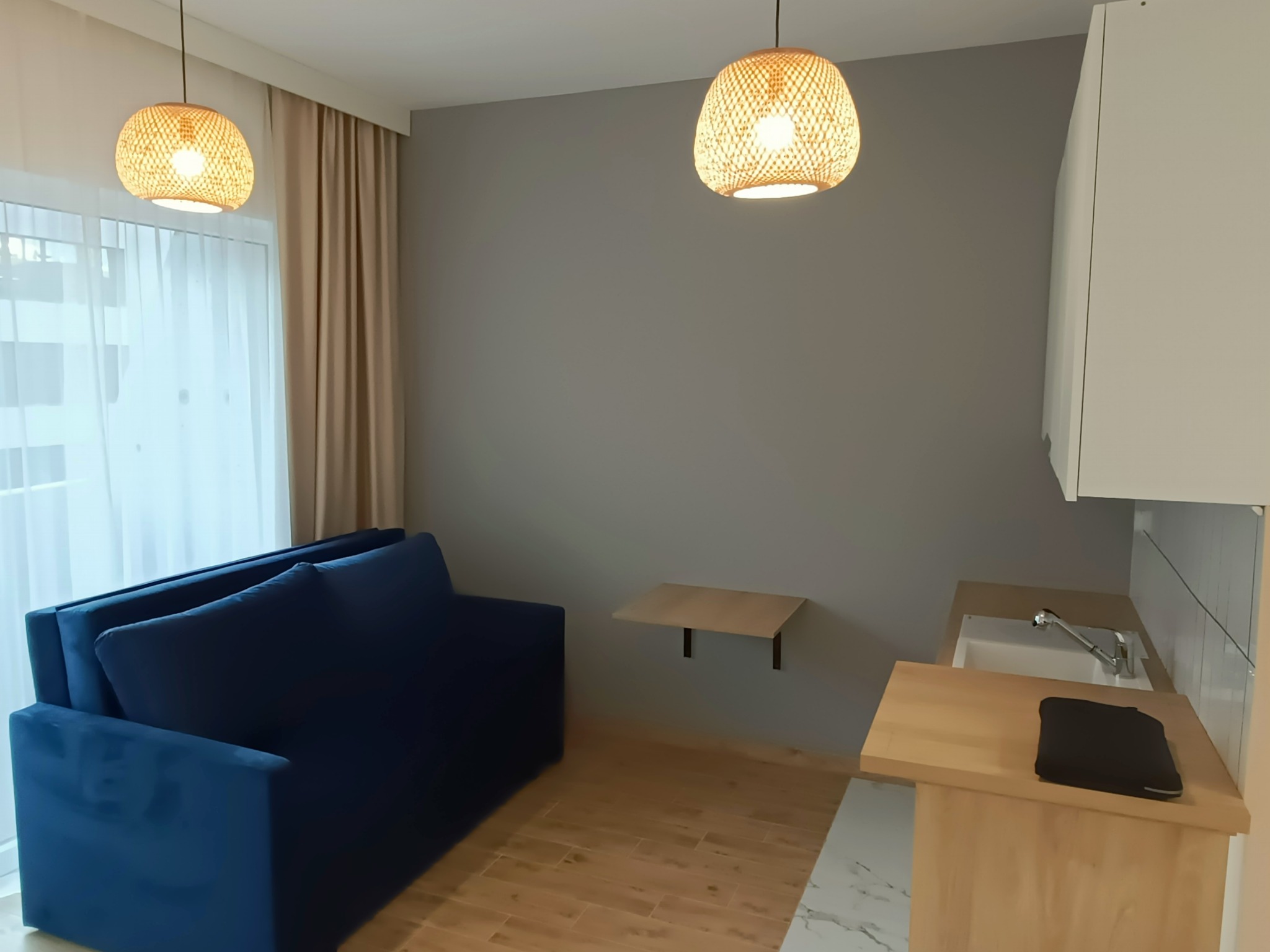 Wnętrze kawalerki po remoncie: granatowa sofa, drewniana podłoga, szara ściana z rozkładanym blatem, aneks kuchenny z jasnym blatem i okrągłe lampy z wikliny.