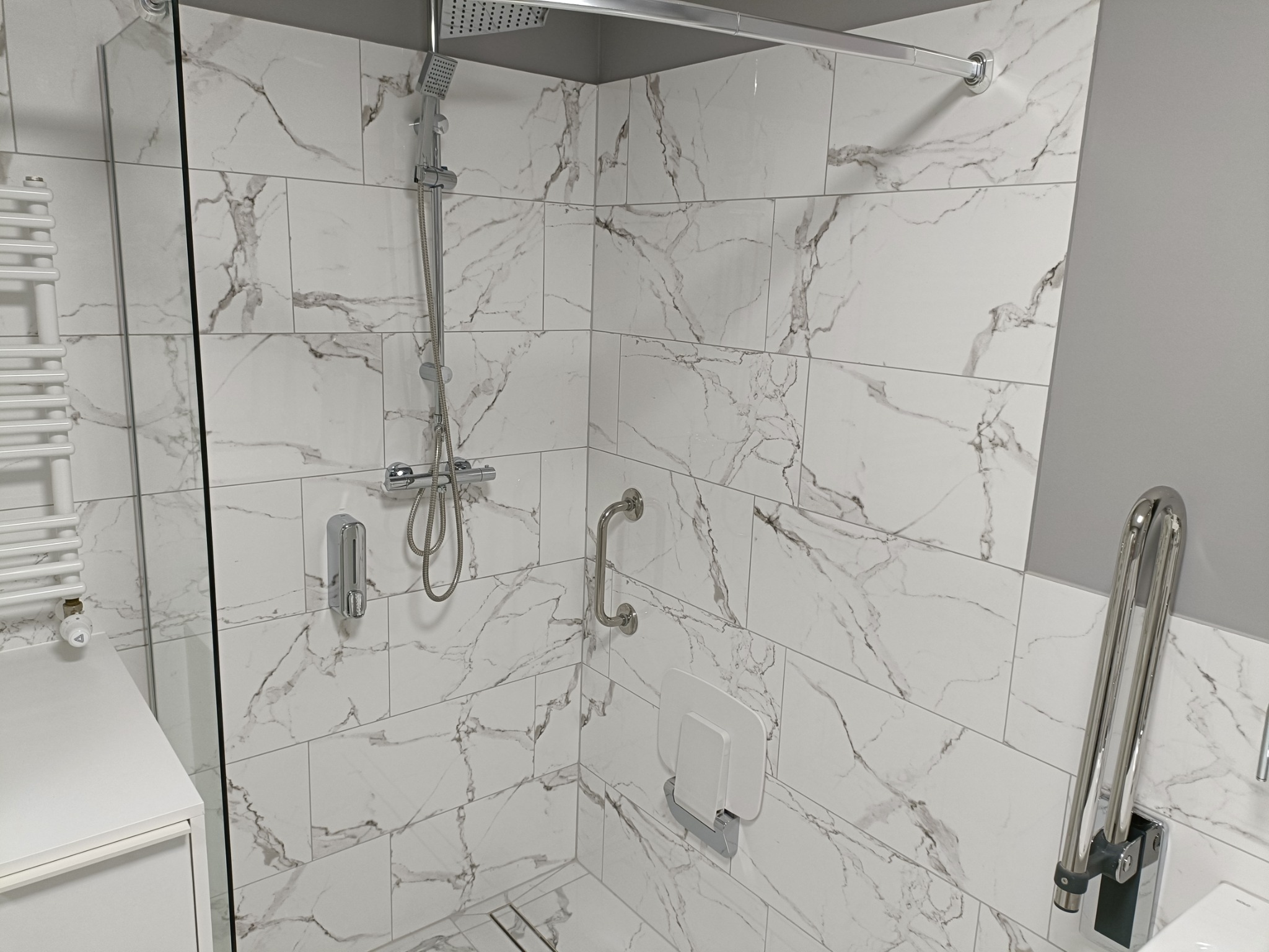 Nowoczesna kabina prysznicowa z marmurowymi płytkami, deszczownicą, szklaną ścianką, siedziskiem i uchwytami dla osób z niepełnosprawnościami.