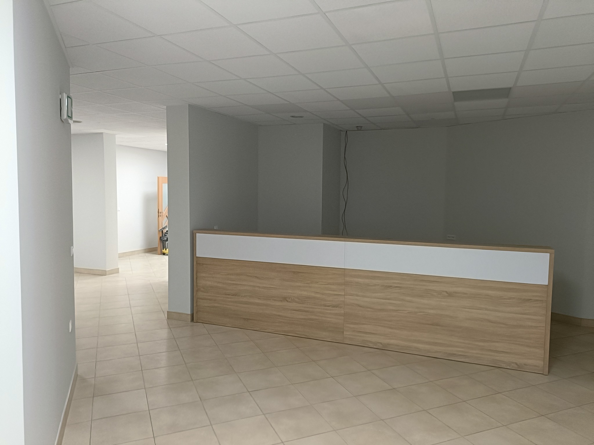 Nowe biuro: jasne płytki podłogowe, białe ściany i sufit kasetonowy, drewniana lada recepcyjna z białym blatem, widok z korytarza.